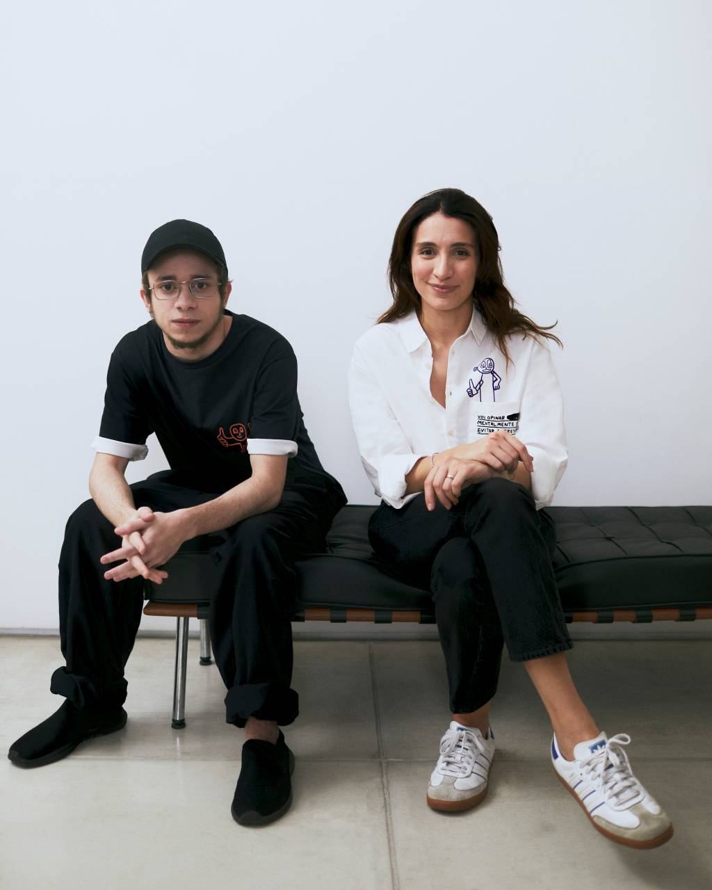 Vanessa Davidowiz e Pedro Vinicio Crédito Pablo Saborido Jovem de preto e boné, moça à direita de cabelos pretos longos vestindo calça preta e blazer branco, ambos sentados em sofá preto