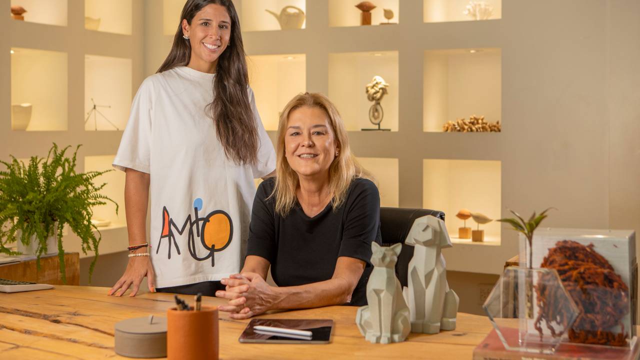 Mãe e filha lado a lado, a filha de pé com camiseta longa branca, a mãe loira de preto sentada â mesa de madeira repleta de cerâmicas