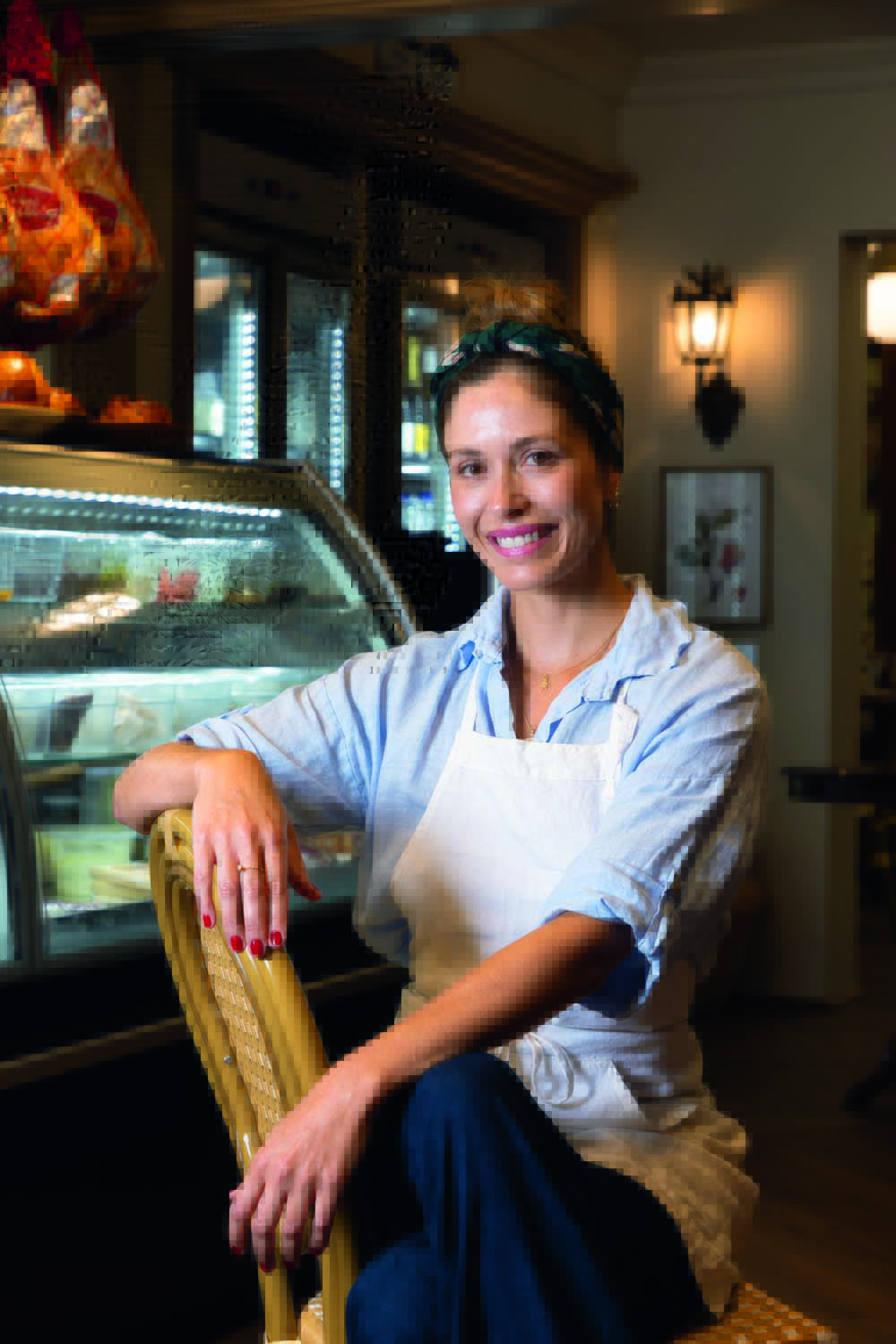 Chef Olivia Maita _ Oli Pane i Salumeria Mulher sentada em cadeira e sorrindo para a foto com vitrine de itens atrás