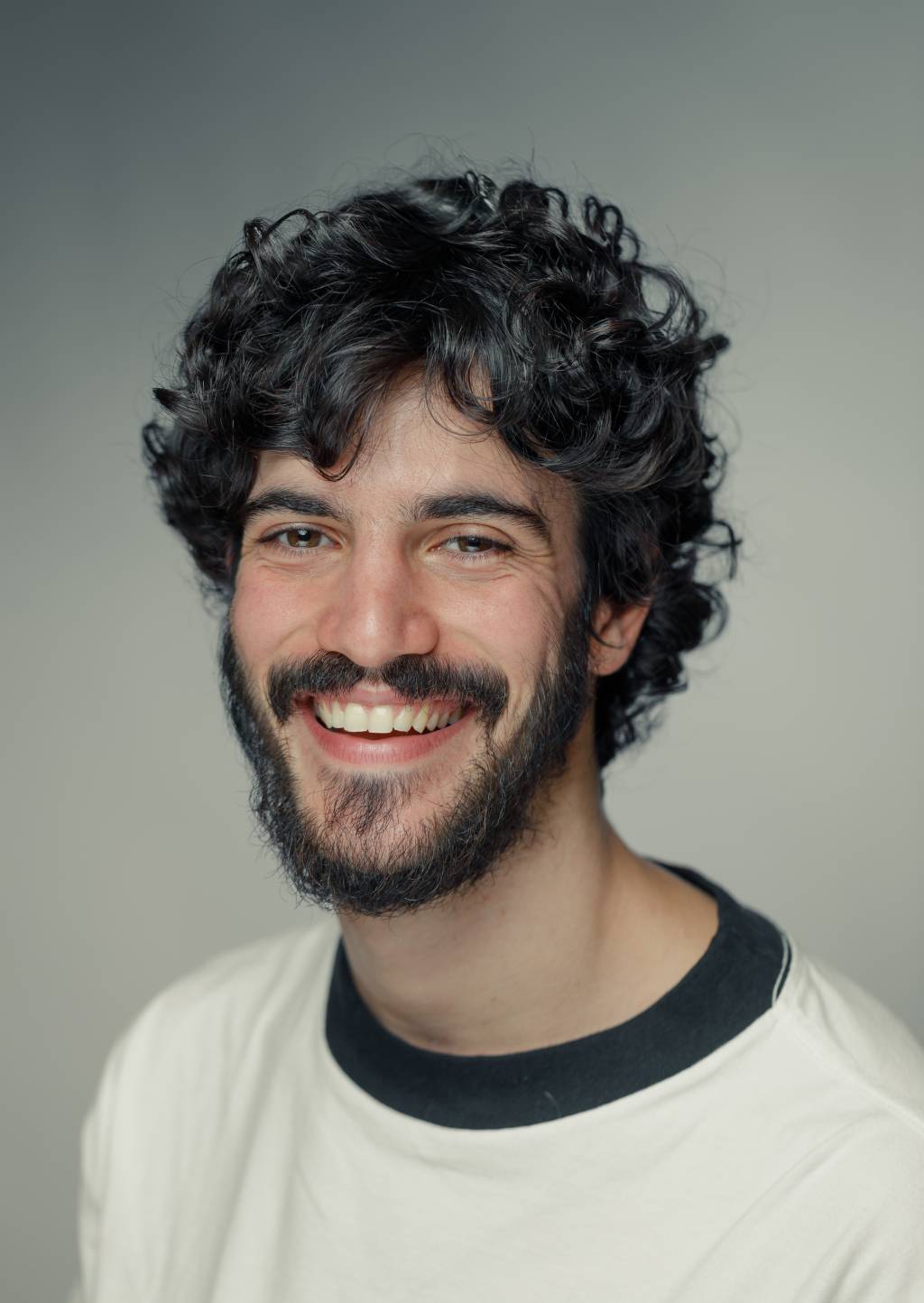 Caio Horowicz Crédito Fabio Audi-Divulgação (1) Moço de cabelo preto encaracolado usa camiseta branca de gola preta e sorri