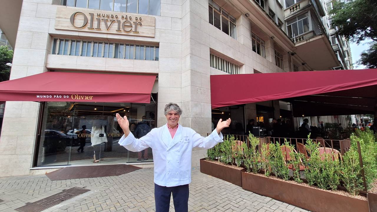 Chef Olivier Anquier, um homem de camisa branca e calça preta em frente à uma padaria