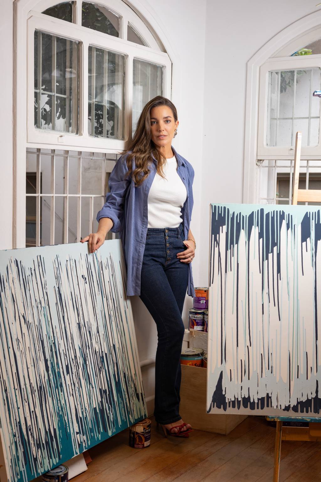 Viviana Ximenes Crédito Leo Martins-VejaSP (1) Artista de cabelos longos castanhos posa ao lado de quadros abstratos em tons de branco e azul