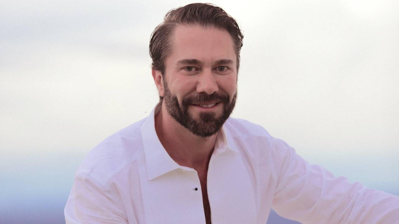 Homem branco de barba sentado em área aberta vestindo peças brancas