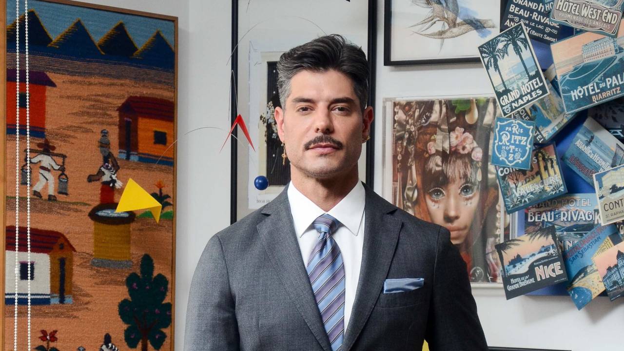 Homem branco com terno cinza em escritório repleto de quadros