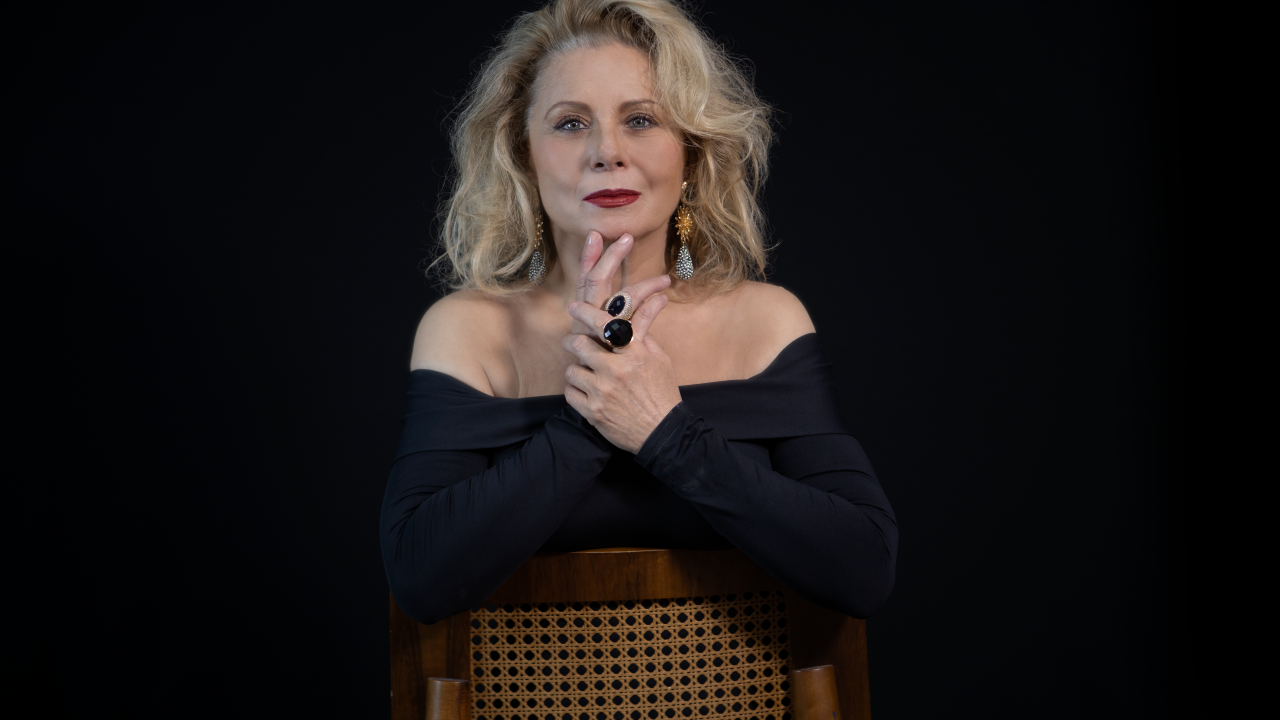 Vera Fischer com as mãos no centro da foto, cadeira à frente