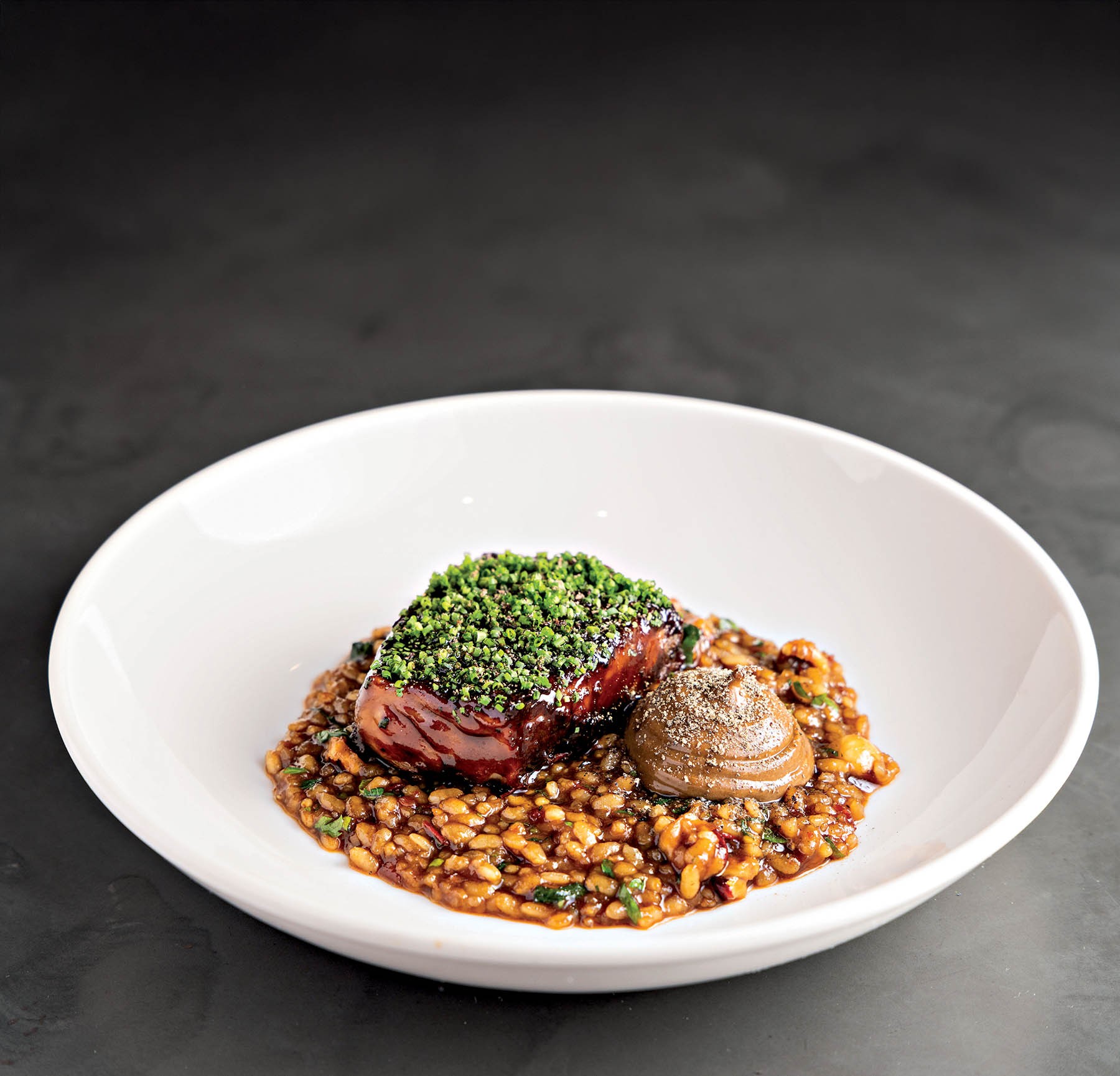 Prato gourmet com carne selada coberta por ervas picadas, servida sobre risoto de gr&atilde;os e um quenelle de pur&ecirc; escuro, em um prato branco fundo