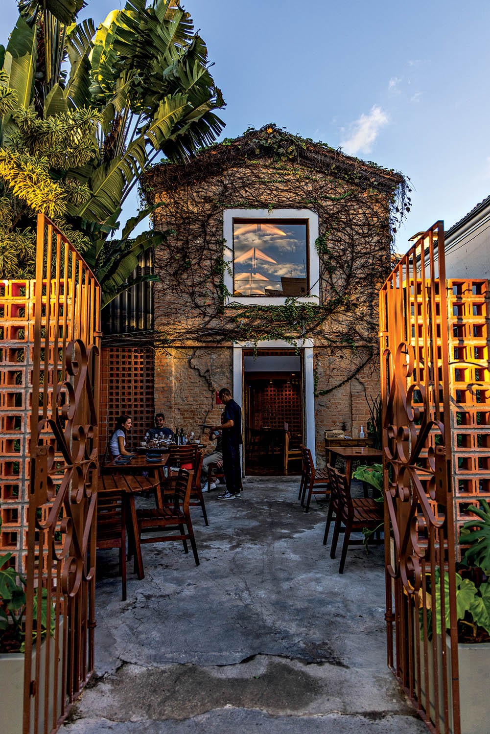 Fachada de tijolos de um restaurante coberta por hera, com um grande port&atilde;o de ferro aberto revelando mesas de madeira no p&aacute;tio. Pessoas sentadas e um gar&ccedil;om atendendo. O c&eacute;u azul com nuvens se reflete na janela superior. Plantas verdes adornam o ambiente