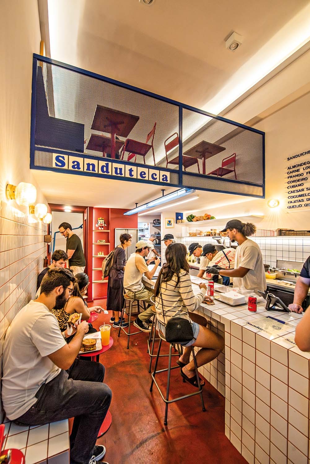 Interior de uma lanchonete moderna com piso vermelho e paredes de azulejos brancos. Clientes comem em mesas e balc&otilde;es, enquanto funcion&aacute;rios preparam lanches. Um letreiro azul com a palavra 