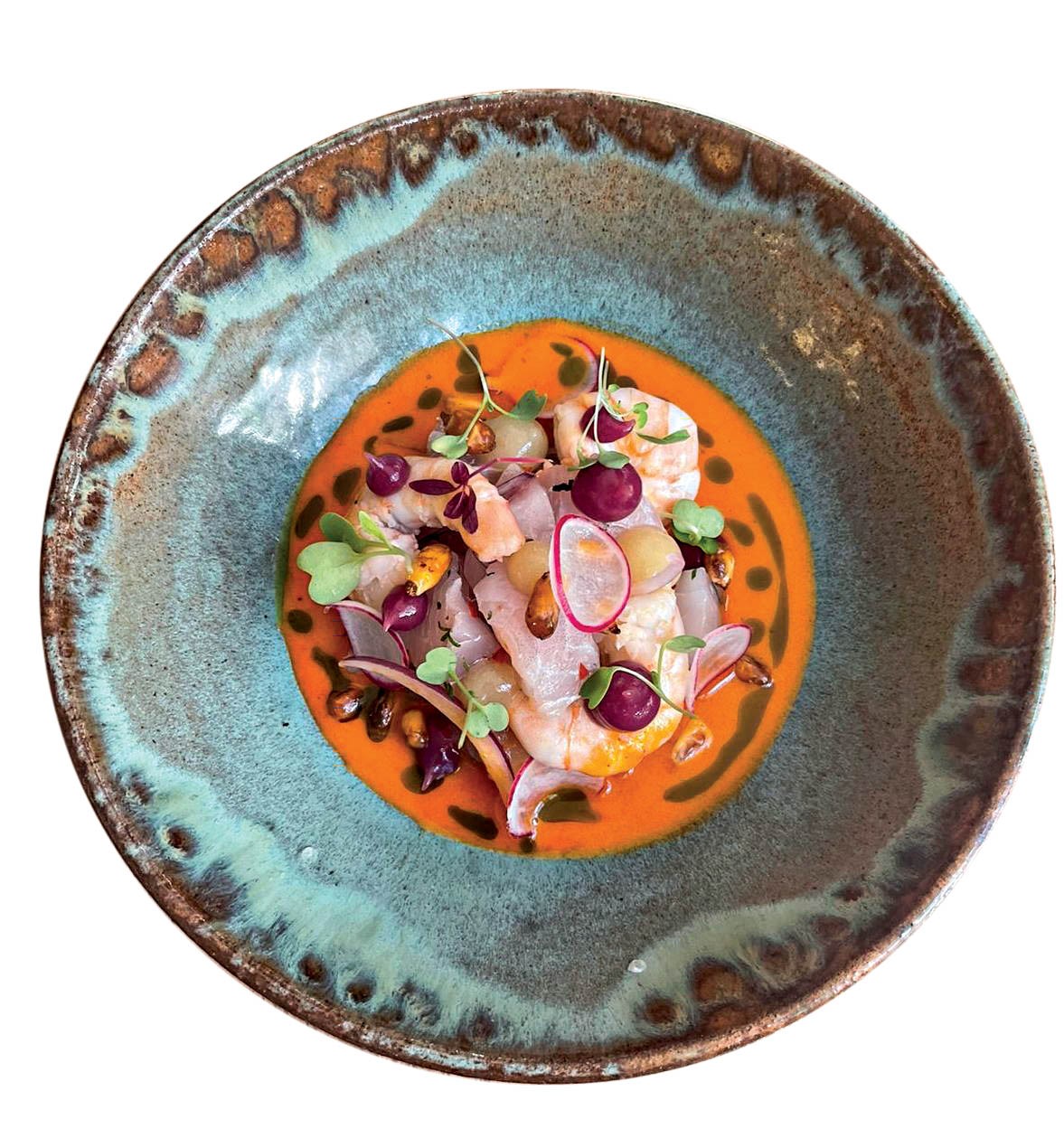 Ceviche colorido em um prato r&uacute;stico azul e marrom, com camar&otilde;es, peixe branco, rabanetes fatiados, uvas roxas e folhas verdes, sobre um molho laranja vibrante