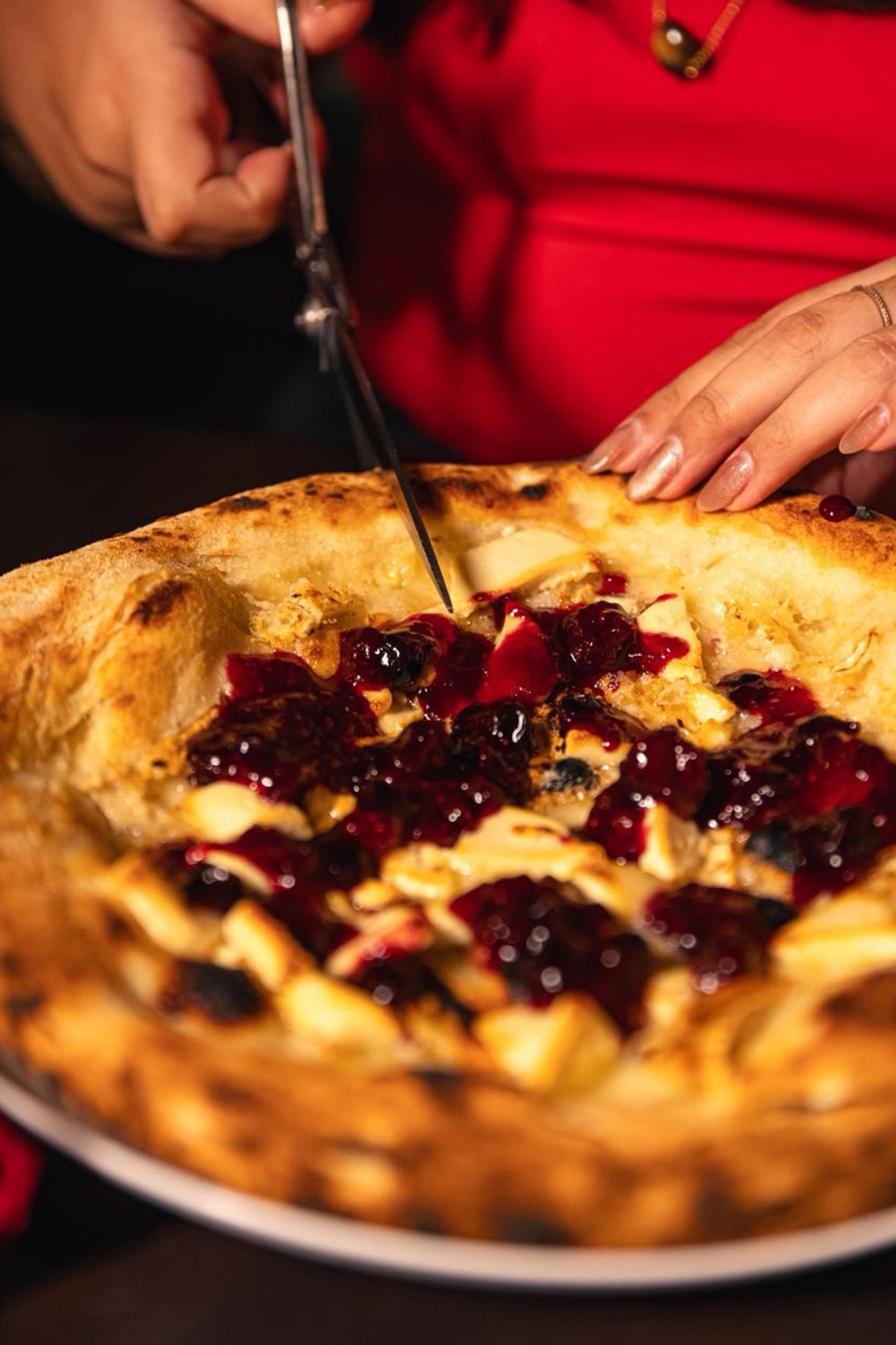M&atilde;os femininas com unhas claras cortando uma pizza doce com massa crocante, coberta por queijo branco e calda escura de frutas vermelhas