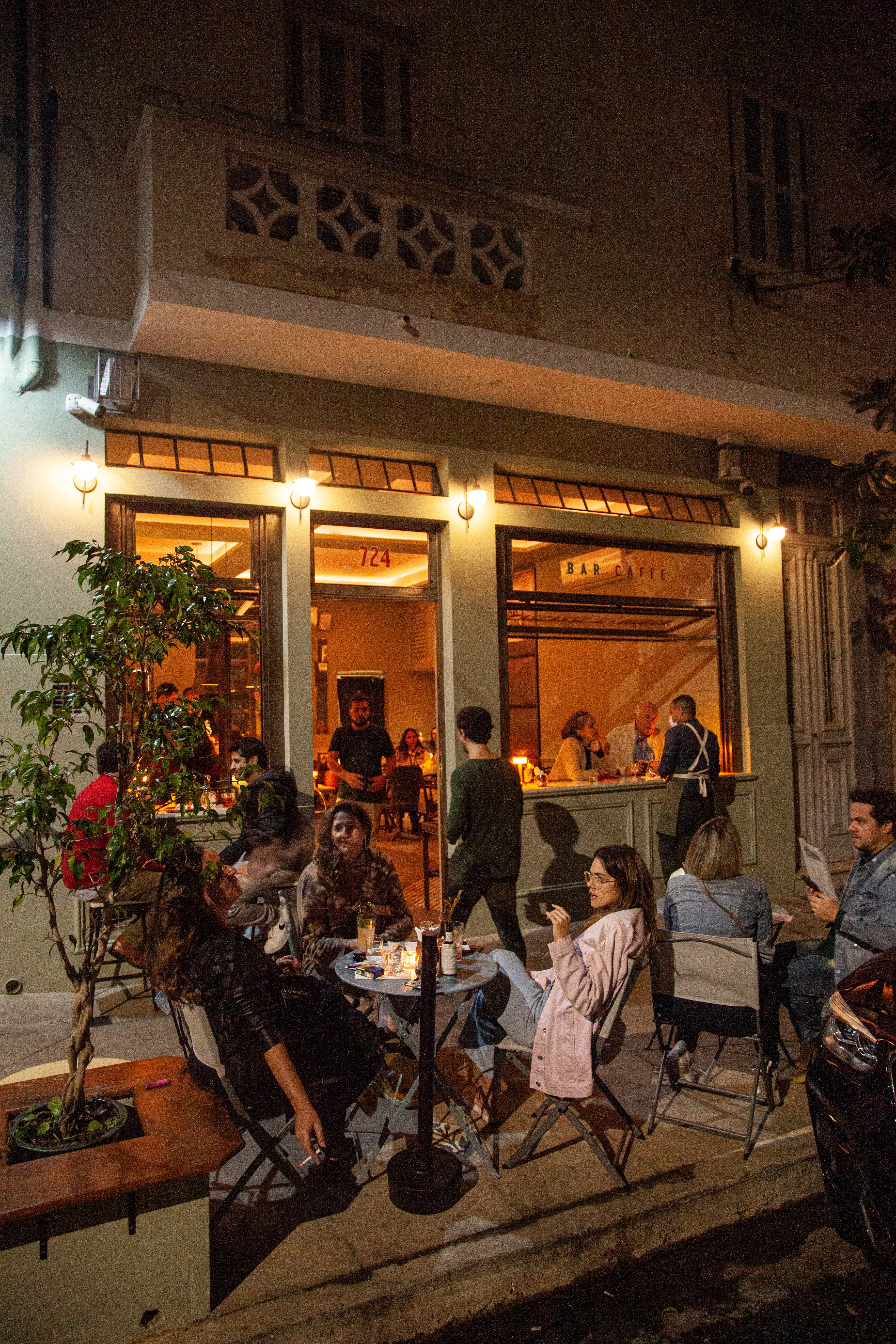 Fachada de bar &agrave; noite com pessoas sentadas em mesas na cal&ccedil;ada e dentro, iluminado por luzes quentes