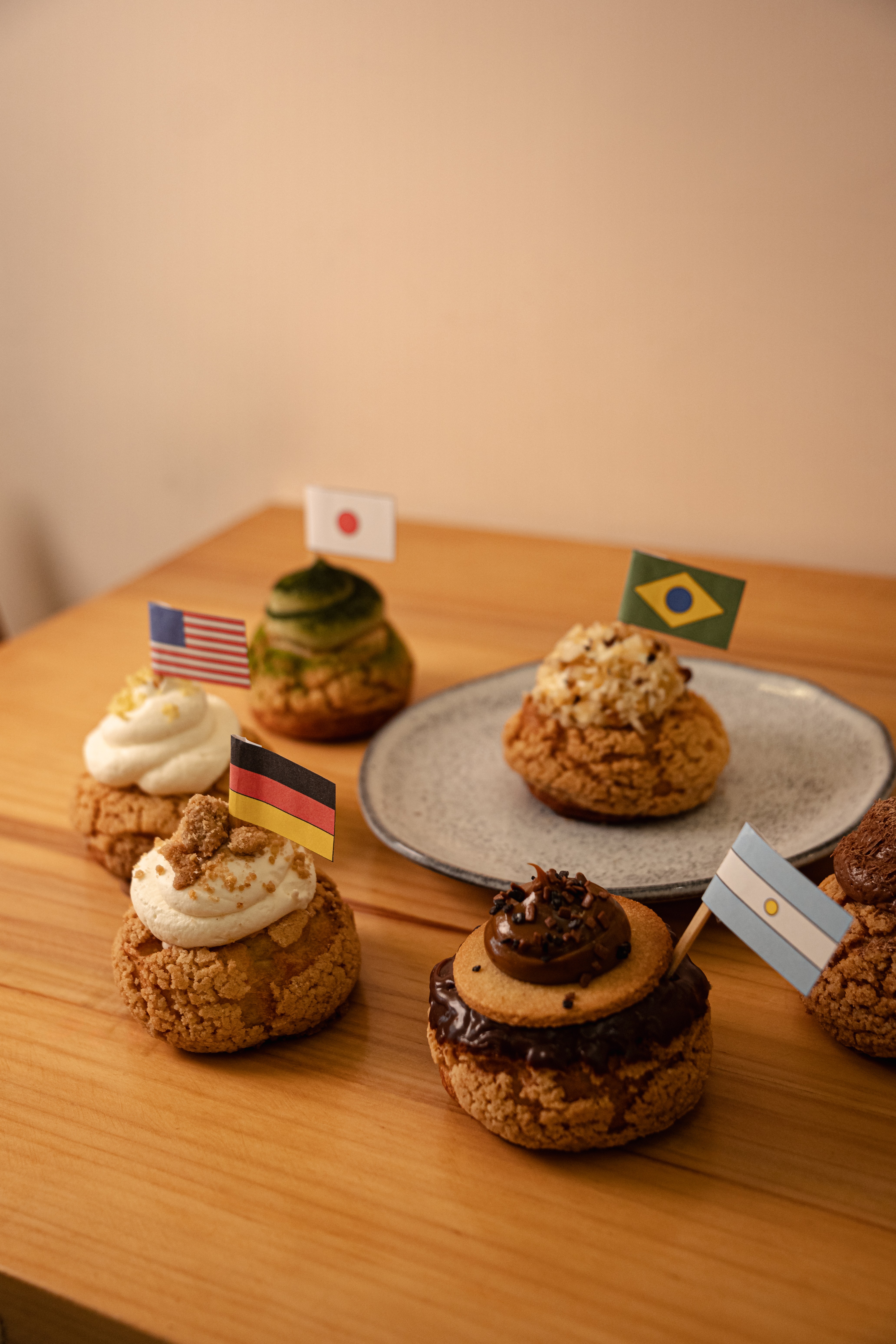 Seis doces redondos com coberturas variadas e pequenas bandeiras de pa&iacute;ses, como Brasil, Jap&atilde;o e EUA, sobre uma mesa de madeira