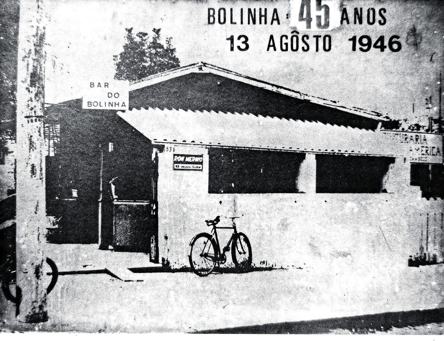 Fachada de um bar simples em preto e branco, com uma bicicleta estacionada na frente. Uma placa indica Bar do Bolinha e outra Dom Merino. No canto superior, texto BOLINHA 45 ANOS 13 AGOSTO 1946