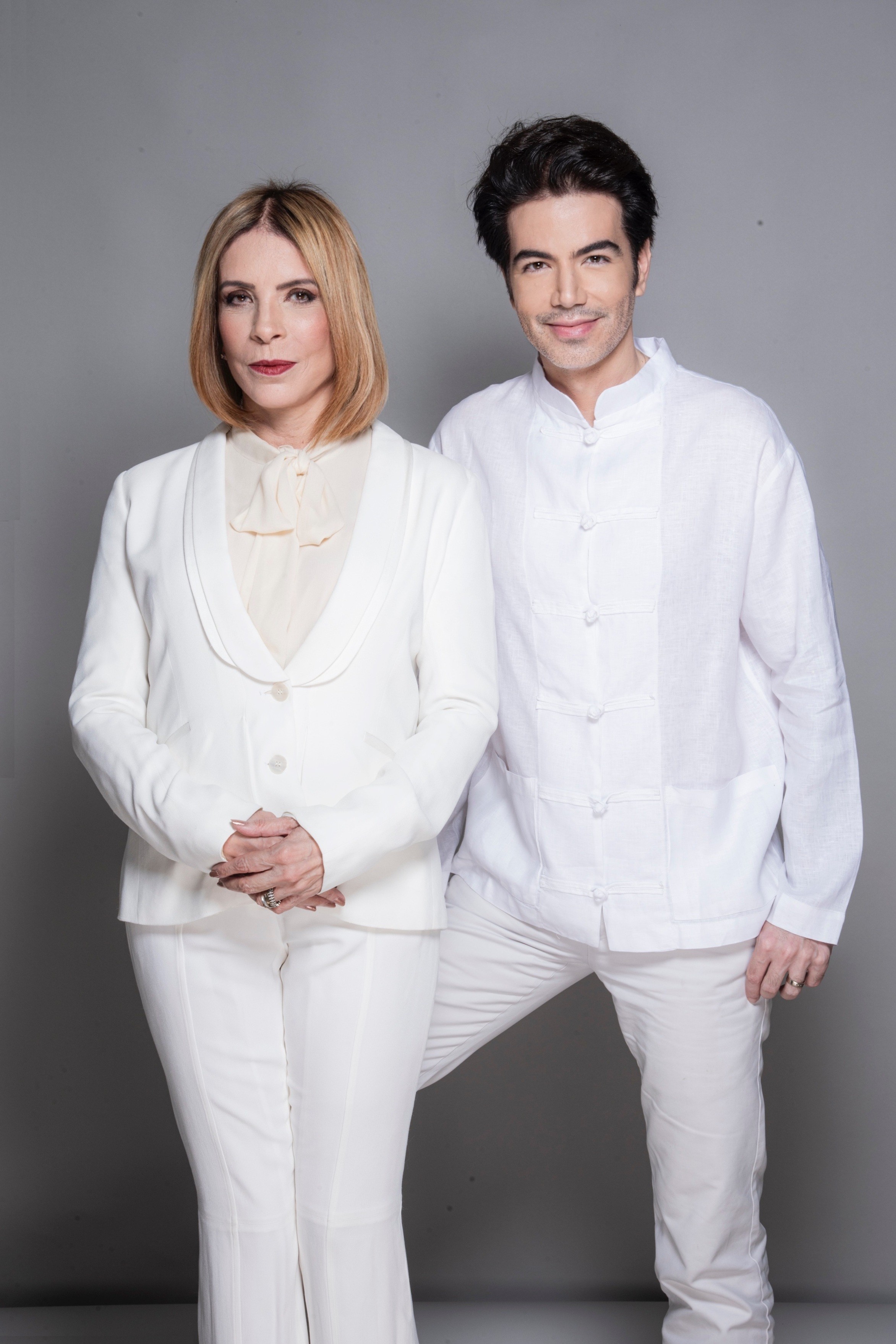 Uma mulher loira de blazer branco e blusa creme com la&ccedil;o, e um homem de camisa branca estilo oriental e cal&ccedil;a branca, posam sorrindo em fundo cinza