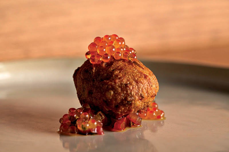 Bolinho frito dourado com cobertura de ovas de salm&atilde;o alaranjadas e peda&ccedil;os de tomate picado, servido em prato cinza com fundo marrom claro