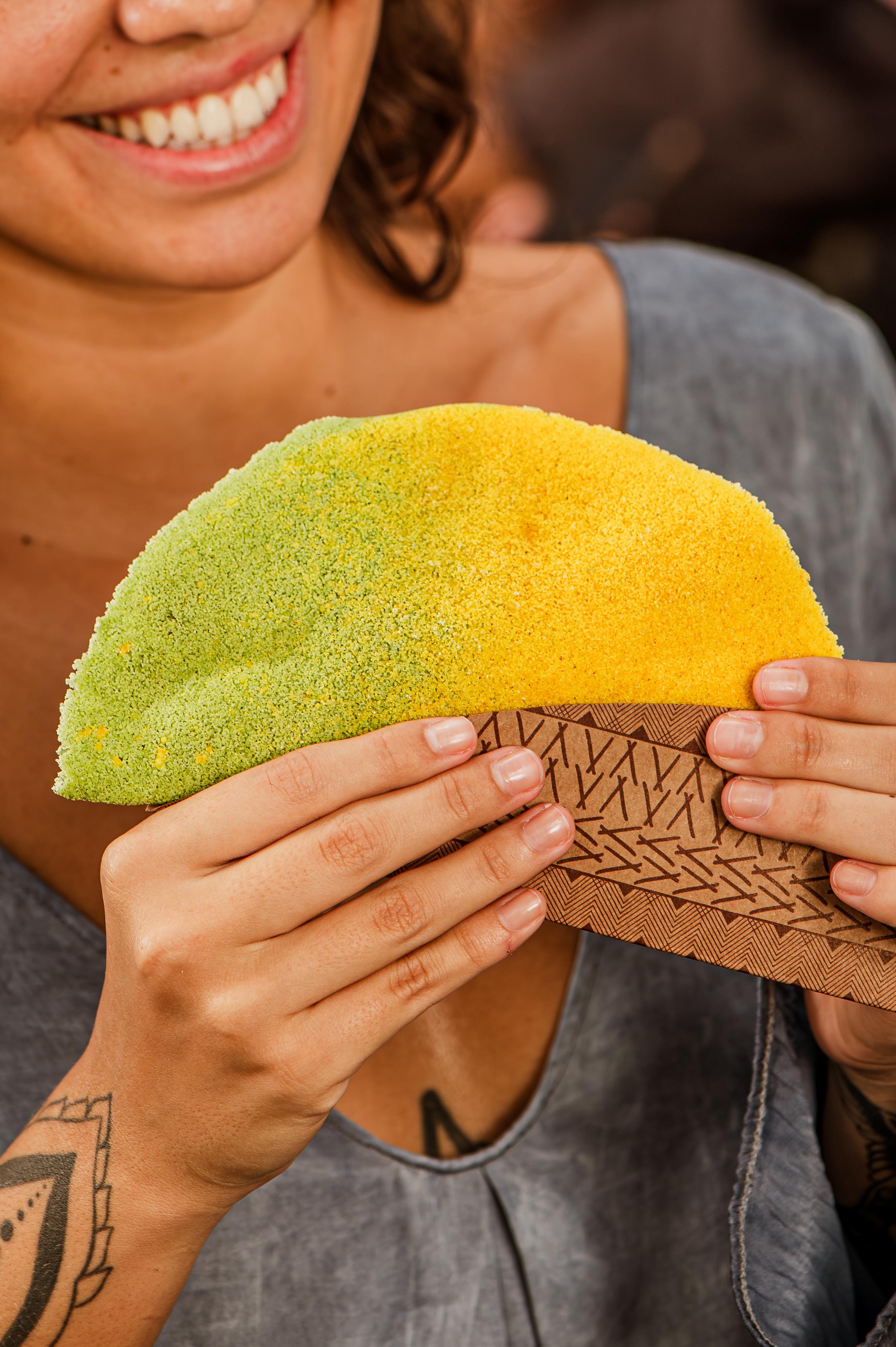 Mulher sorridente segurando uma tapioca colorida, metade verde e metade amarela, com as m&atilde;os em um suporte de papel marrom estampado