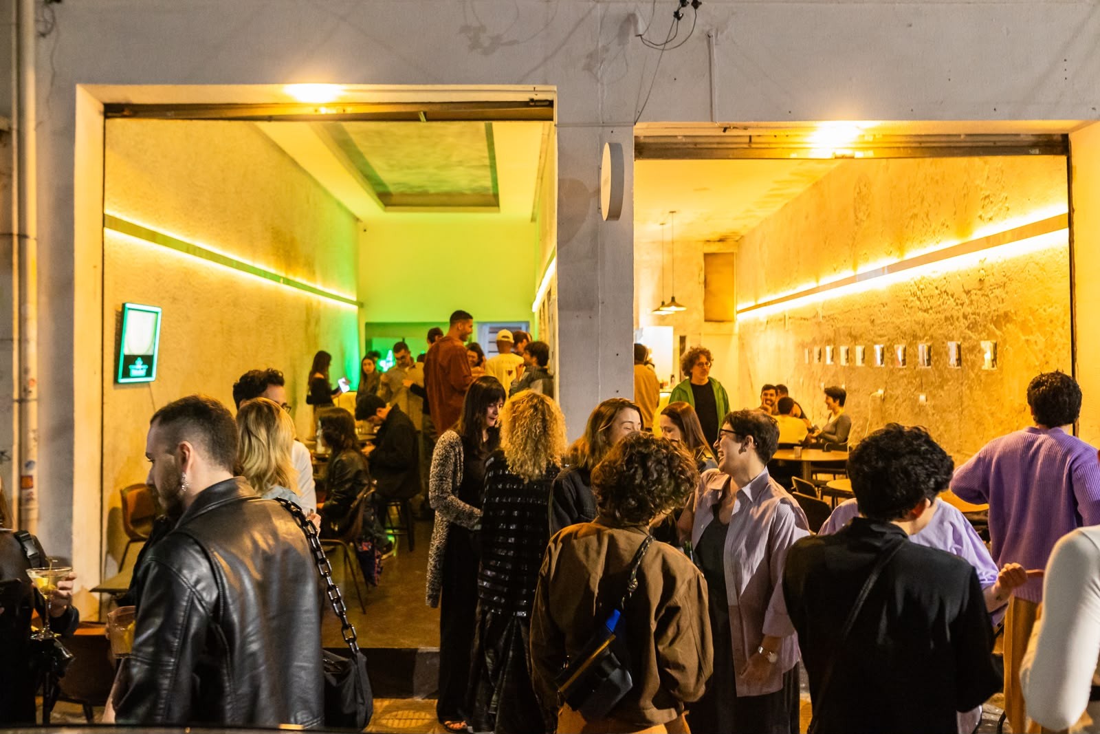 Pessoas conversando e socializando em um bar ou evento noturno, com ilumina&ccedil;&atilde;o amarela e verde neon nas paredes, criando uma atmosfera vibrante