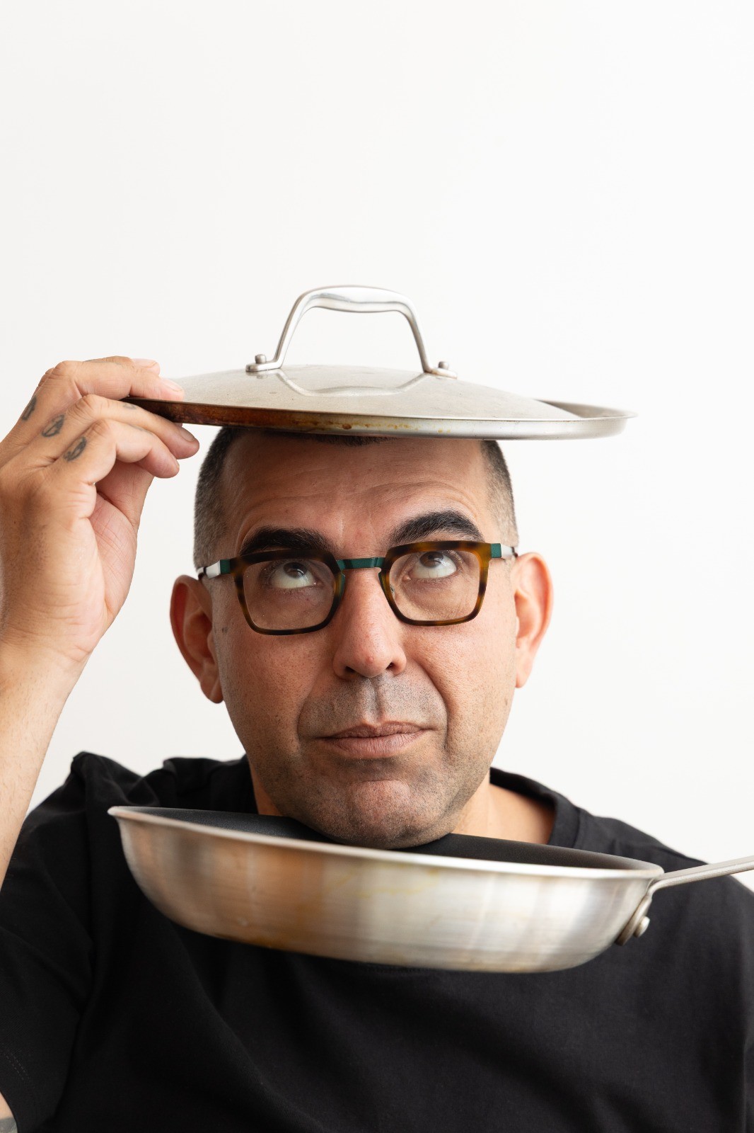 Homem de &oacute;culos e camiseta preta, com express&atilde;o s&eacute;ria, equilibra uma tampa de panela na cabe&ccedil;a e uma frigideira no pesco&ccedil;o, olhando para cima