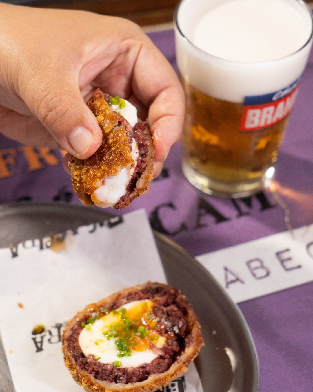 M&atilde;o segura um bolinho de carne frito com recheio branco, enquanto outro bolinho com ovo cozido e cheiro-verde repousa sobre papel. Ao fundo, um copo de cerveja e um tecido roxo com letras.
