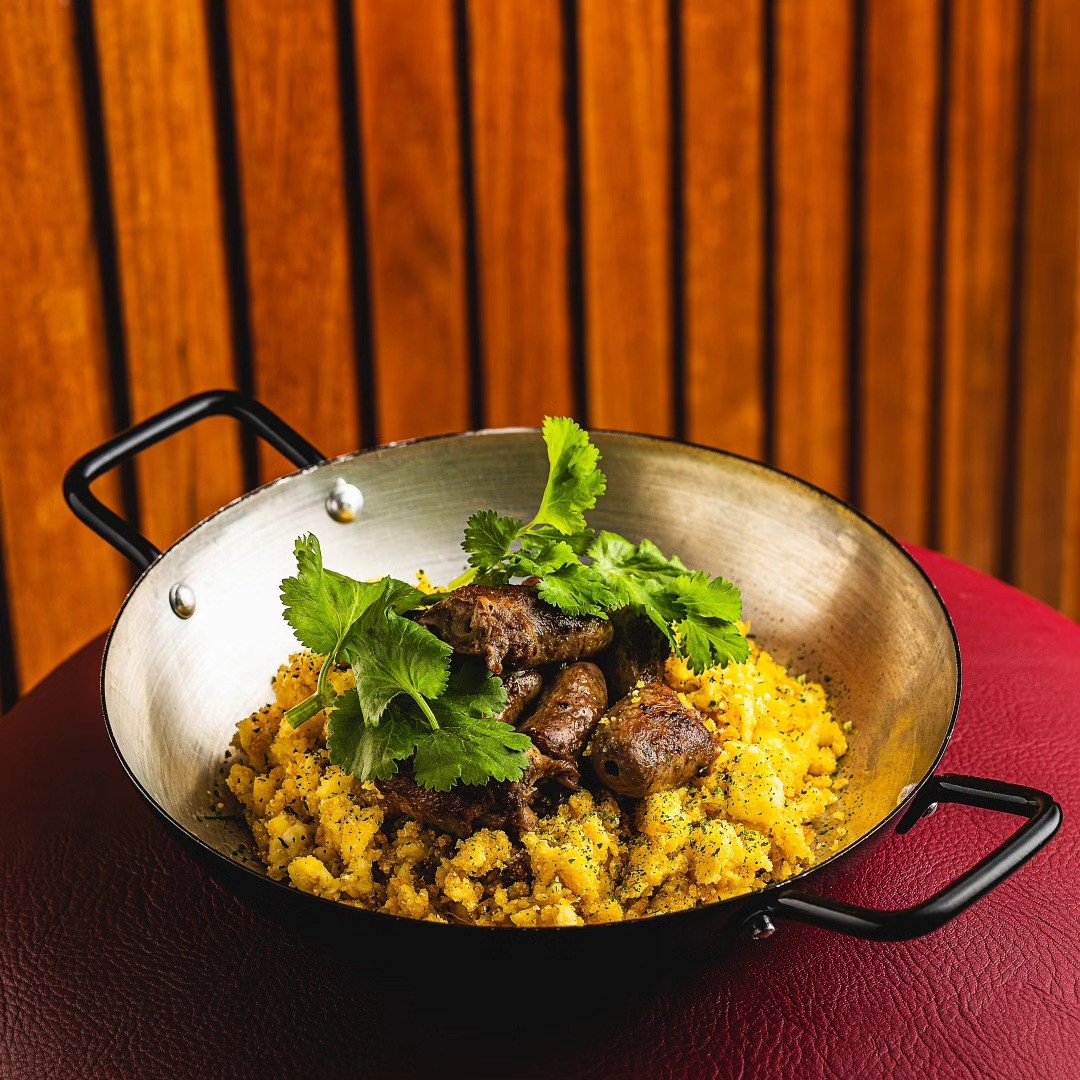 Por&ccedil;&atilde;o de carne de sol com farofa amarela e folhas de coentro fresco, servida em frigideira de metal com al&ccedil;as pretas sobre superf&iacute;cie vermelha, com fundo de madeira vertical