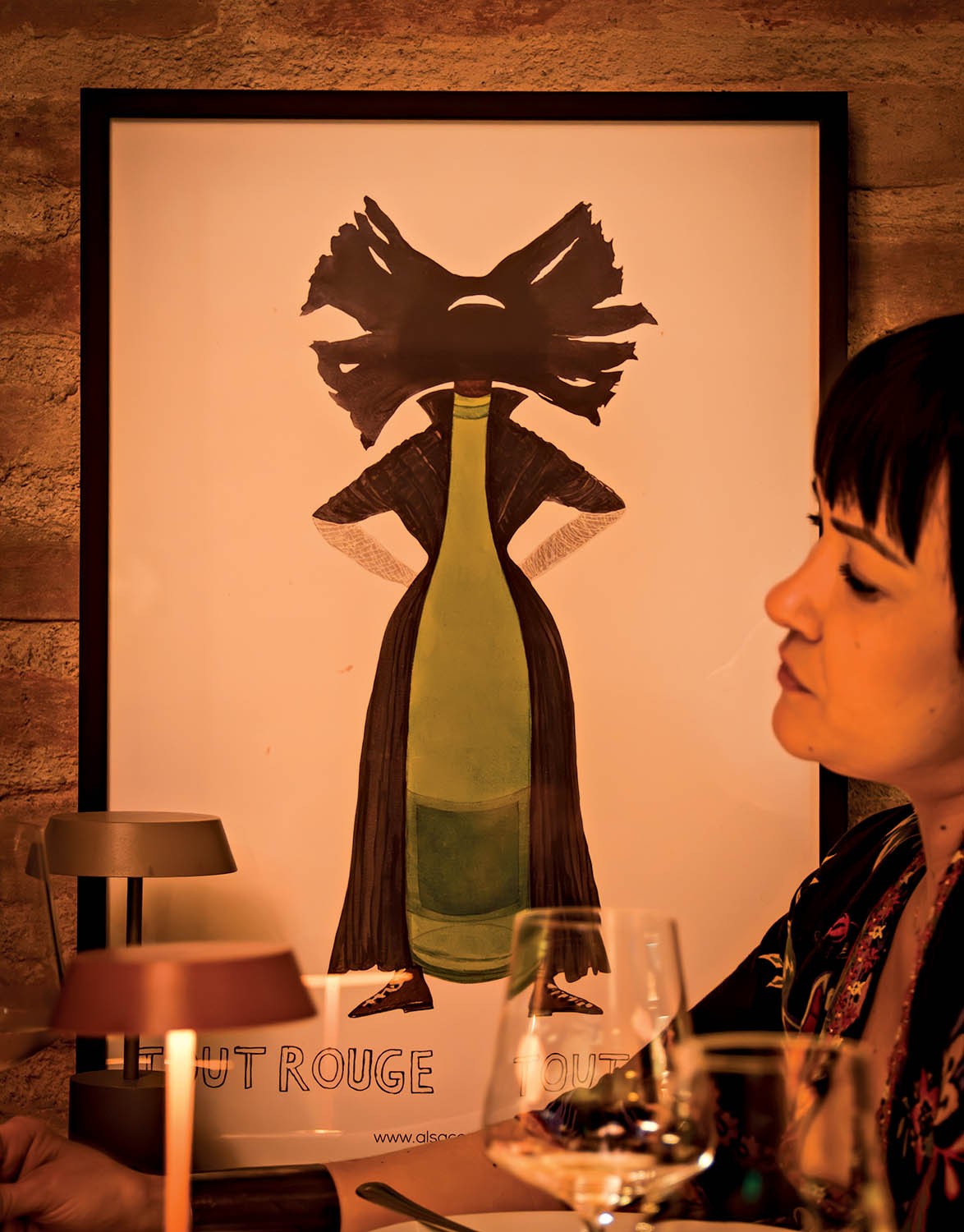 Mulher de perfil, com cabelo escuro e franja, sentada &agrave; mesa com ta&ccedil;as de vinho e uma lumin&aacute;ria. Ao fundo, um p&ocirc;ster com uma garrafa de vinho verde vestindo um casaco escuro e sapatos, com uma cabe&ccedil;a abstrata preta e texto CHUT ROUGE e www.alsace.com