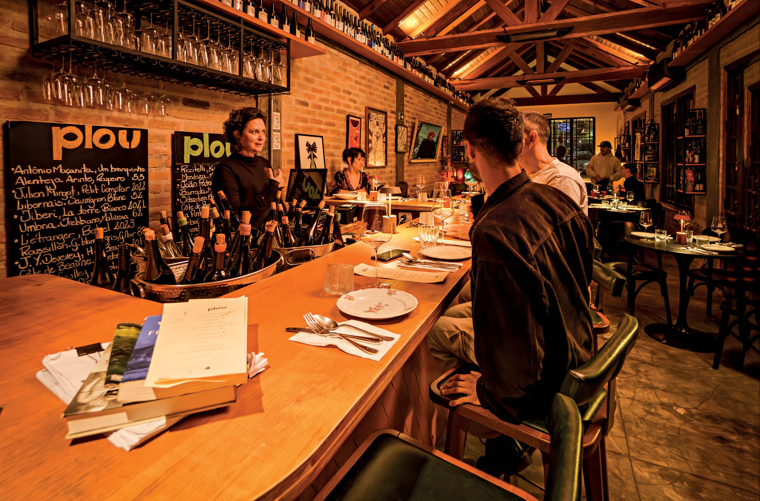 Mulher de cabelo cacheado escuro e blusa preta atende clientes em um bar de madeira com garrafas de vinho e quadros de giz com menus. Clientes sentados em bancos altos e mesas ao fundo, sob ilumina&ccedil;&atilde;o quente e teto de madeira.