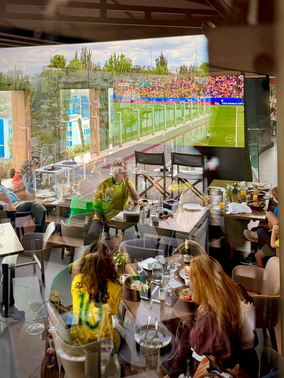 Pessoas assistem a um jogo de futebol em um tel&atilde;o gigante, refletido em uma janela de um restaurante. Uma mulher de camisa amarela com o n&uacute;mero 10 est&aacute; sentada de costas, enquanto outra pessoa de camisa amarela e verde observa o tel&atilde;o. Mesas com pratos e bebidas est&atilde;o dispostas no ambiente.