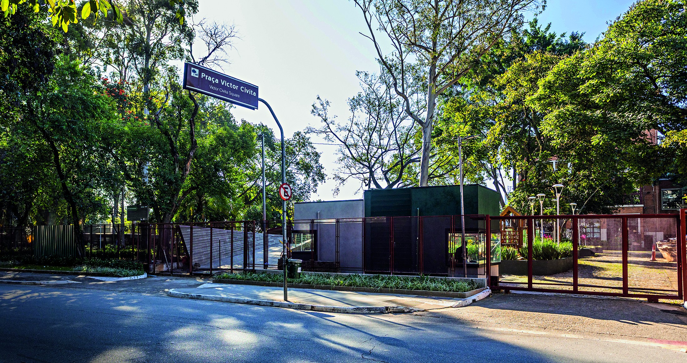 Placa azul com Praça Victor Civita em poste metálico, cercada por árvores frondosas e portões de madeira e metal. Ao fundo, construções baixas e um parque com gramado e brinquedos. O sol ilumina a cena, criando sombras no asfalto.