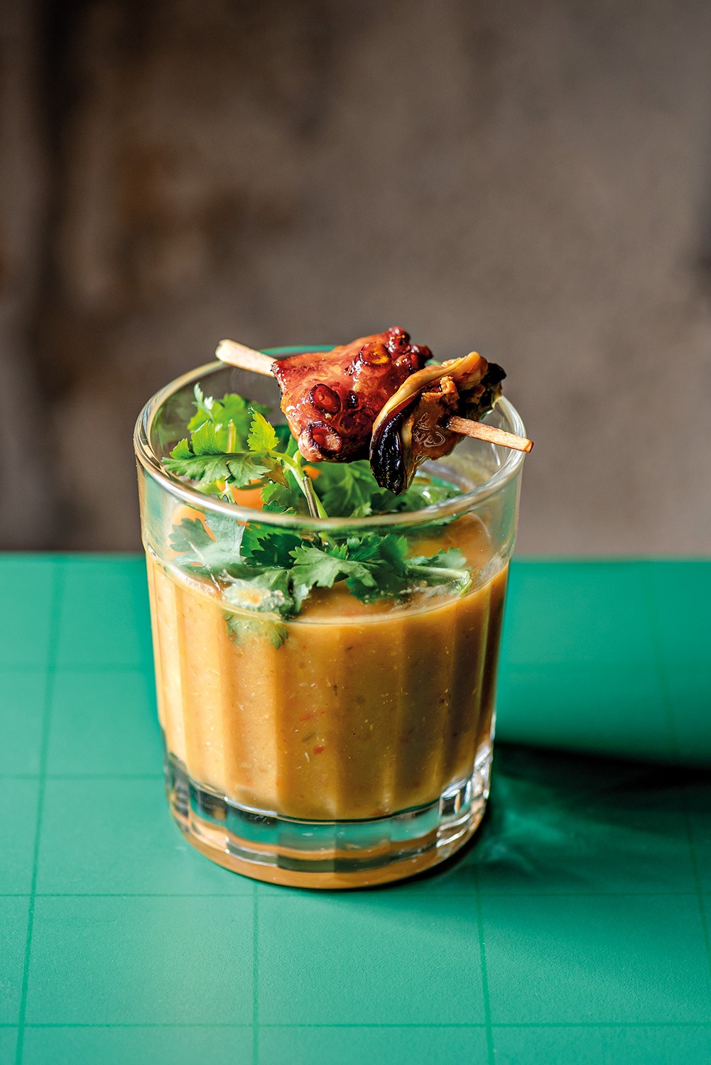 Copo de vidro com caldo cremoso alaranjado, decorado com folhas de coentro e dois espetinhos de frutos do mar grelhados, um de polvo e outro de mexilh&atilde;o. O copo est&aacute; sobre uma superf&iacute;cie verde-&aacute;gua quadriculada, com fundo cinza desfocado