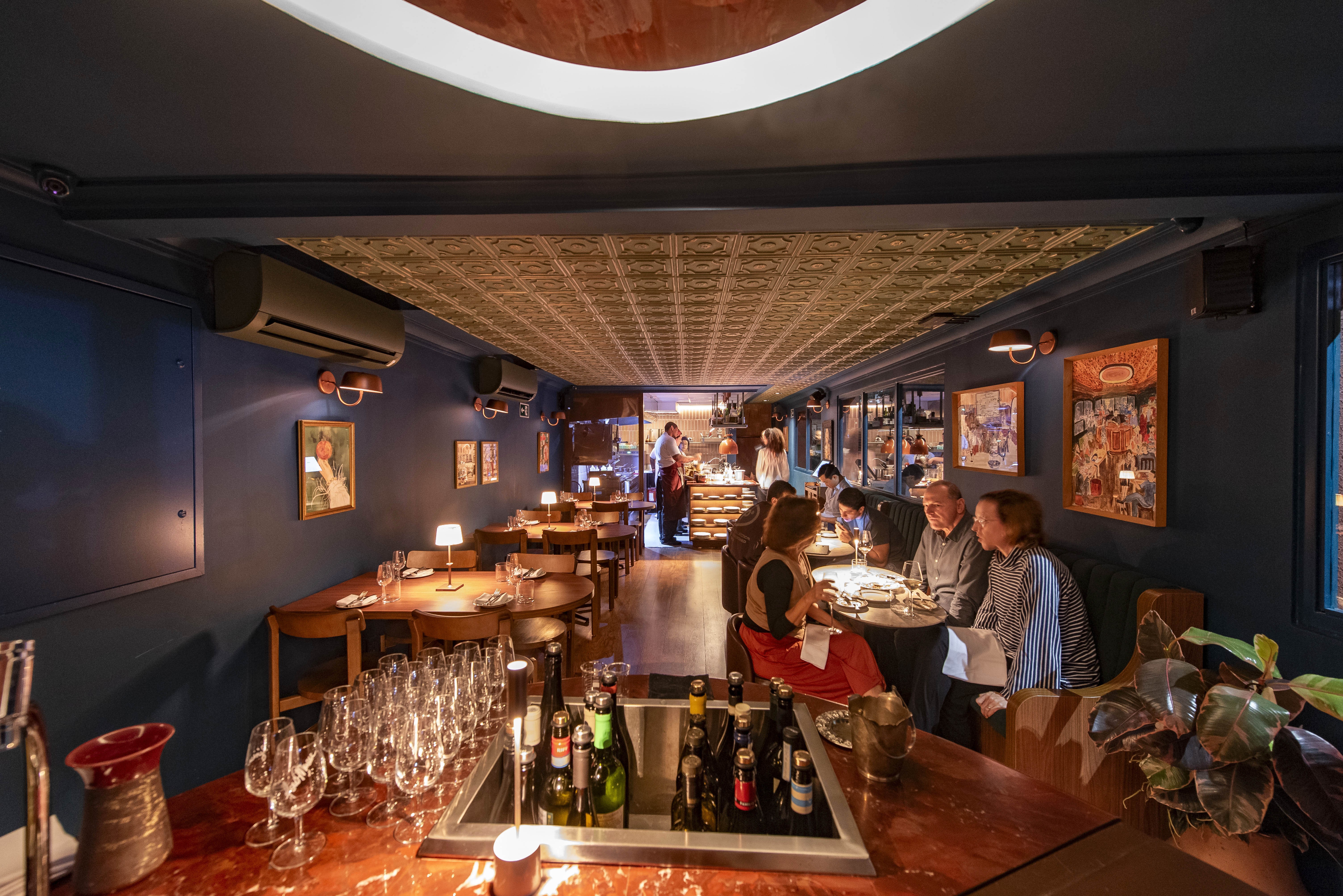 Interior de restaurante com teto dourado texturizado e paredes azul-marinho. Clientes sentados em mesas de madeira escuras, alguns conversando e outros olhando para seus pratos. Um bar com garrafas de vinho e ta&ccedil;as vazias ocupa o primeiro plano.