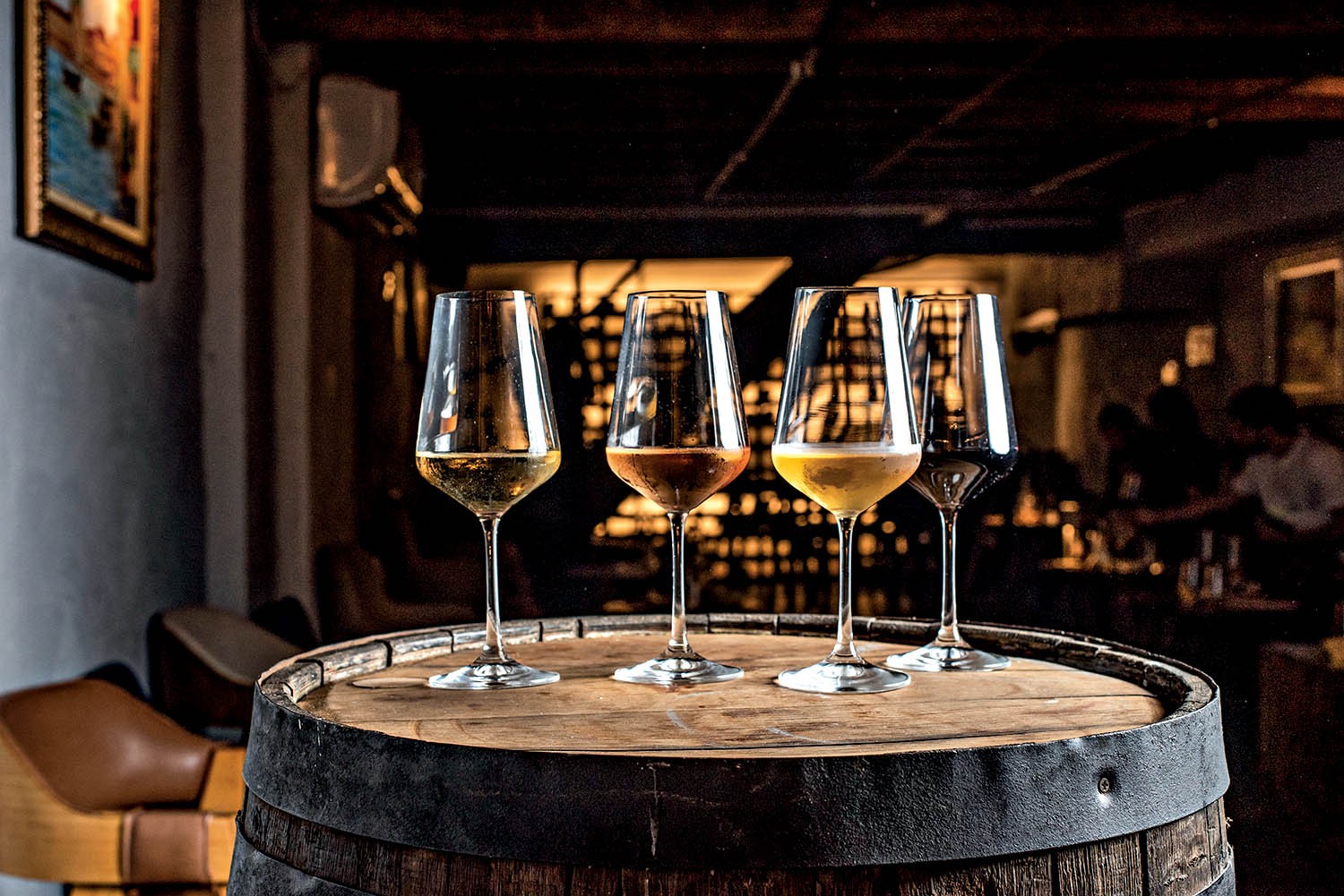 Quatro ta&ccedil;as de vinho com diferentes tonalidades de l&iacute;quido, do claro ao escuro, sobre um barril de madeira em um ambiente escuro de bar com pessoas desfocadas ao fundo