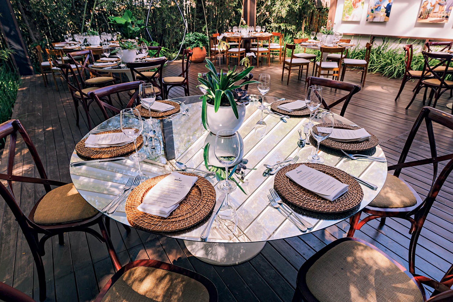 Mesa redonda de vidro com oito lugares, posta para refei&ccedil;&atilde;o ao ar livre. Cada lugar tem um sousplat de palha, guardanapo branco com menu, talheres e ta&ccedil;as. Um vaso branco com planta verde centraliza a mesa. O ch&atilde;o &eacute; de madeira e h&aacute; outras mesas ao fundo, cercadas por vegeta&ccedil;&atilde;o
