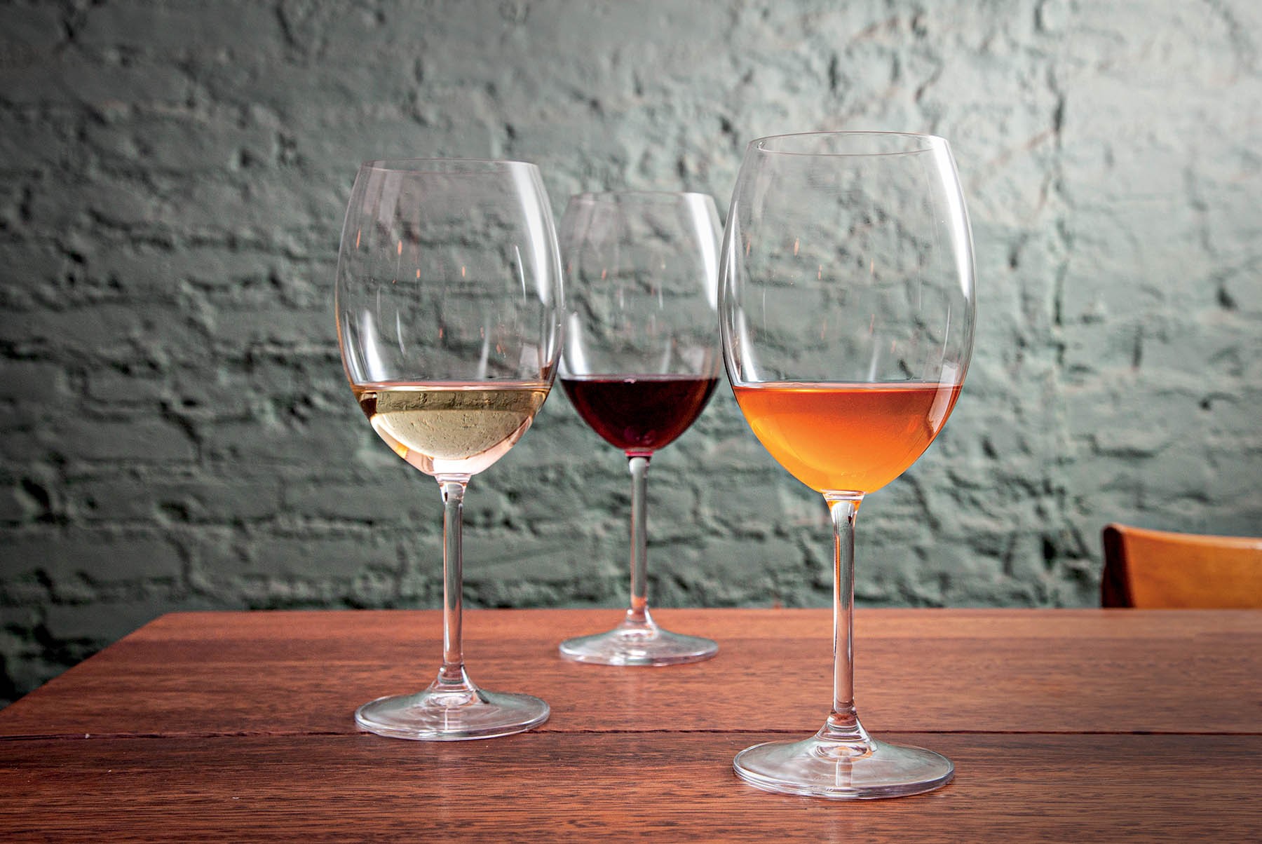 Tr&ecirc;s ta&ccedil;as de vinho em uma mesa de madeira escura, com uma parede de tijolos cinza ao fundo. Da esquerda para a direita, as ta&ccedil;as cont&ecirc;m vinho branco, tinto e ros&eacute;.