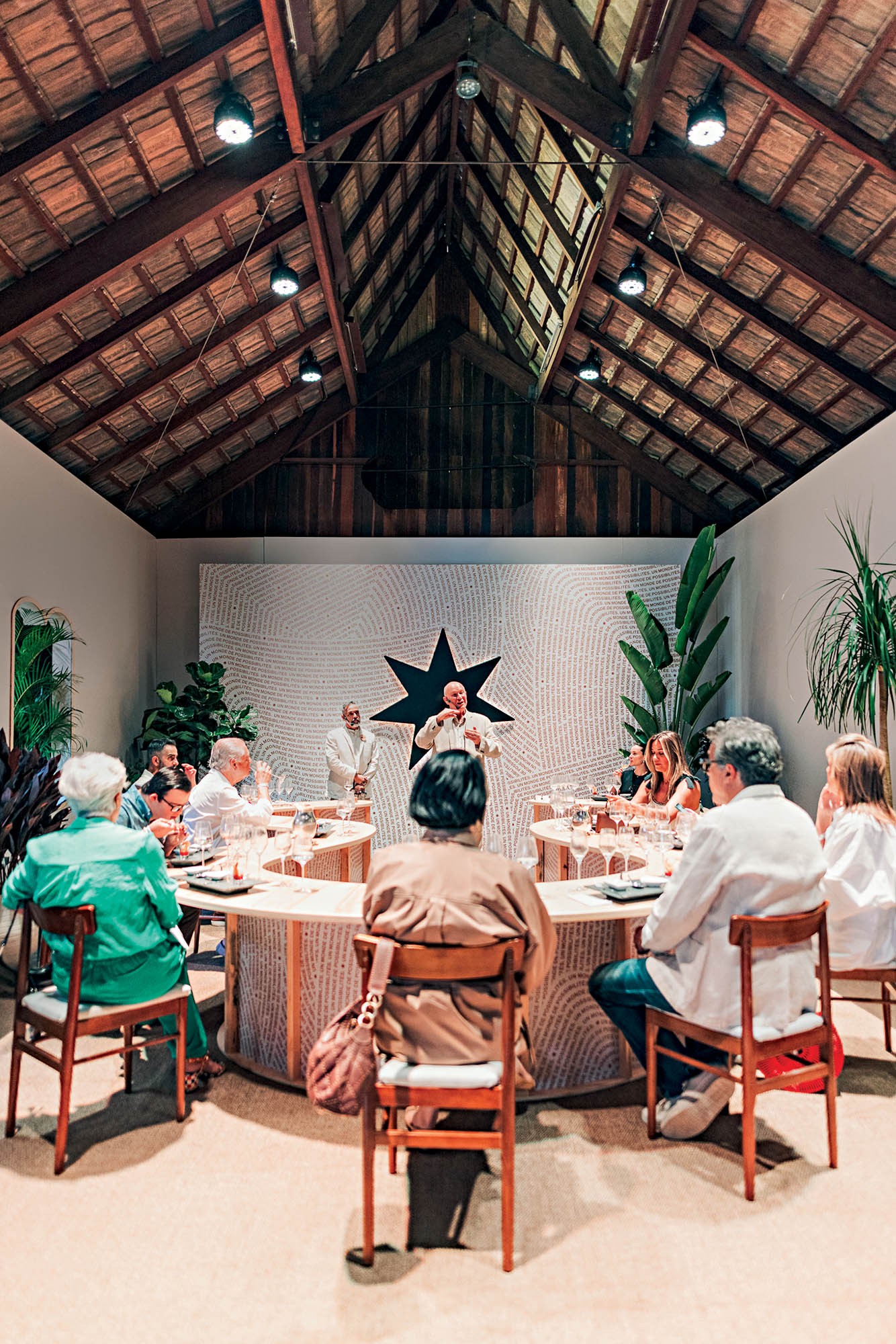 Pessoas sentadas em mesas redondas, assistindo a um homem de camisa branca falando em frente a uma parede branca com uma estrela preta e plantas verdes, sob um teto de madeira com vigas expostas