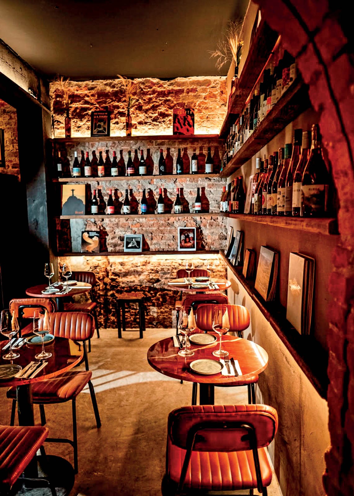 Interior de um restaurante ou bar com mesas redondas de madeira e cadeiras de couro marrom, dispostas em um ambiente aconchegante com paredes de tijolos aparentes e prateleiras cheias de garrafas de vinho iluminadas por luzes quentes