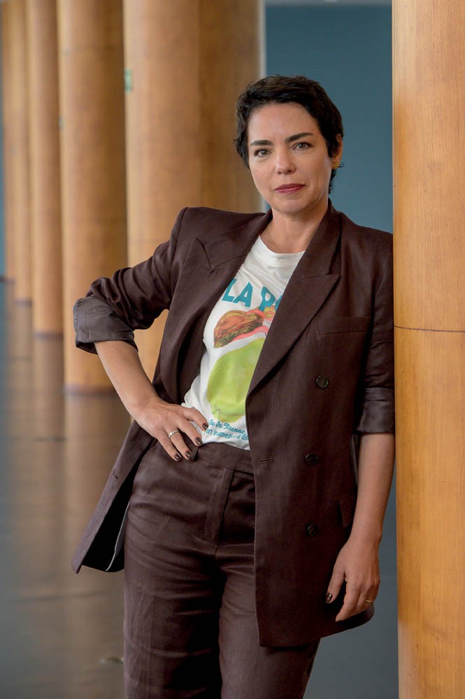 Mulher de pele clara e cabelos curtos escuros, vestindo blazer e cal&ccedil;a marrons, com camiseta branca estampada, posa com a m&atilde;o na cintura e o bra&ccedil;o esquerdo apoiado em uma coluna de madeira clara, olhando para frente com express&atilde;o s&eacute;ria