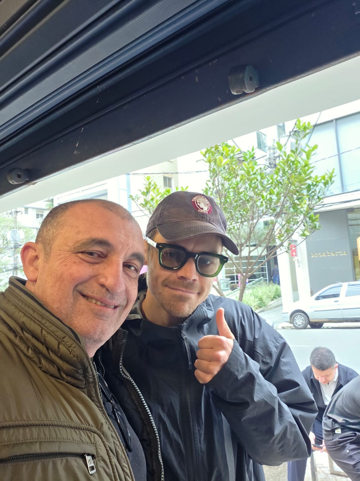 Dois homens sorrindo para a c&acirc;mera, um com jaqueta verde-oliva e outro de bon&eacute;, &oacute;culos e jaqueta preta, fazendo sinal de positivo. Ao fundo, uma rua com &aacute;rvores e um carro branco estacionado
