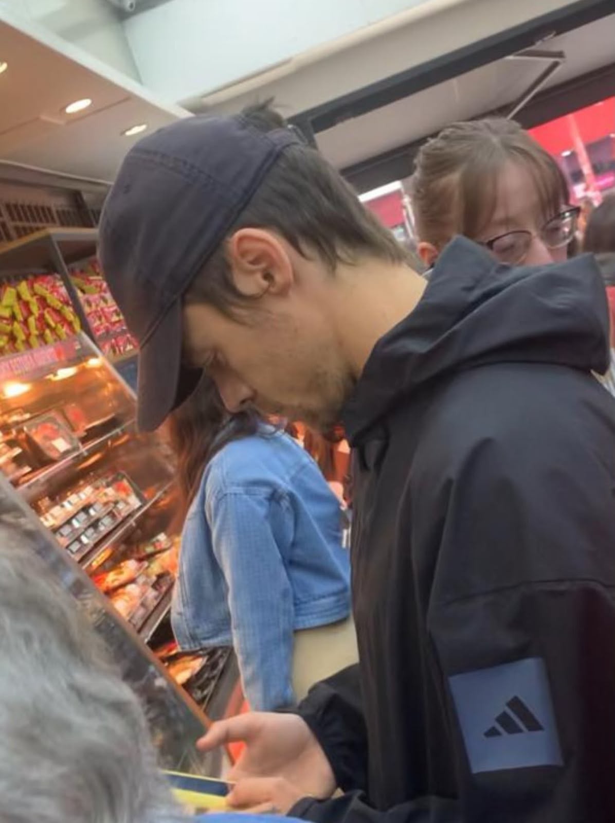 Homem de bon&eacute; azul-marinho e jaqueta preta com logo Adidas no bra&ccedil;o, olhando para baixo em um supermercado. Ao fundo, uma mulher de jaqueta jeans e outra de &oacute;culos. Prateleiras com produtos iluminados &agrave; esquerda.