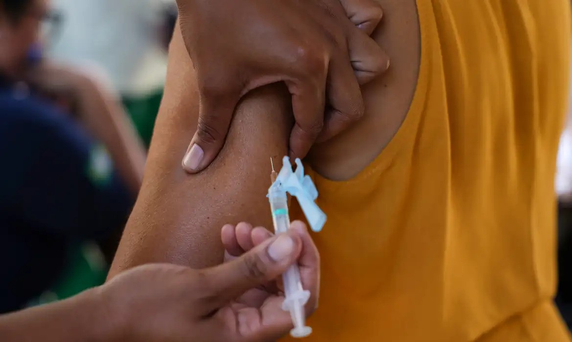 Uma pessoa de pele escura, vestindo blusa amarela, recebendo uma injeção no braço esquerdo. Uma mão segura a seringa com agulha fina, enquanto outra pinça a pele do braço.