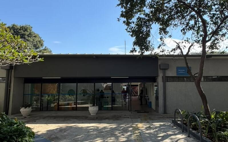 Fachada de um prédio moderno com paredes claras e uma grande entrada de vidro, sob um céu azul. Há plantas verdes em vasos brancos e um bicicletário à direita, com árvores frondosas sombreando parte do chão.