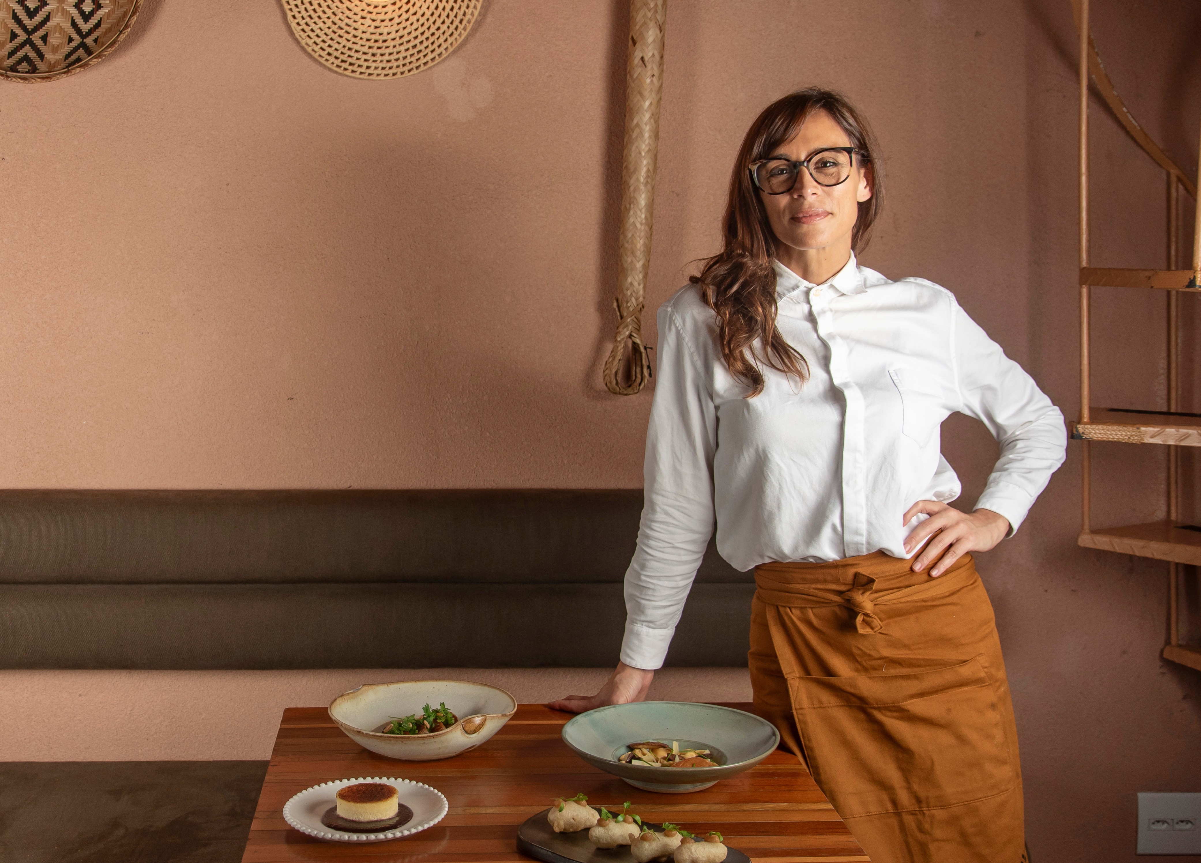 Mulher de &oacute;culos, camisa branca e saia marrom, com a m&atilde;o na cintura, posa em restaurante com pratos de comida sobre a mesa
