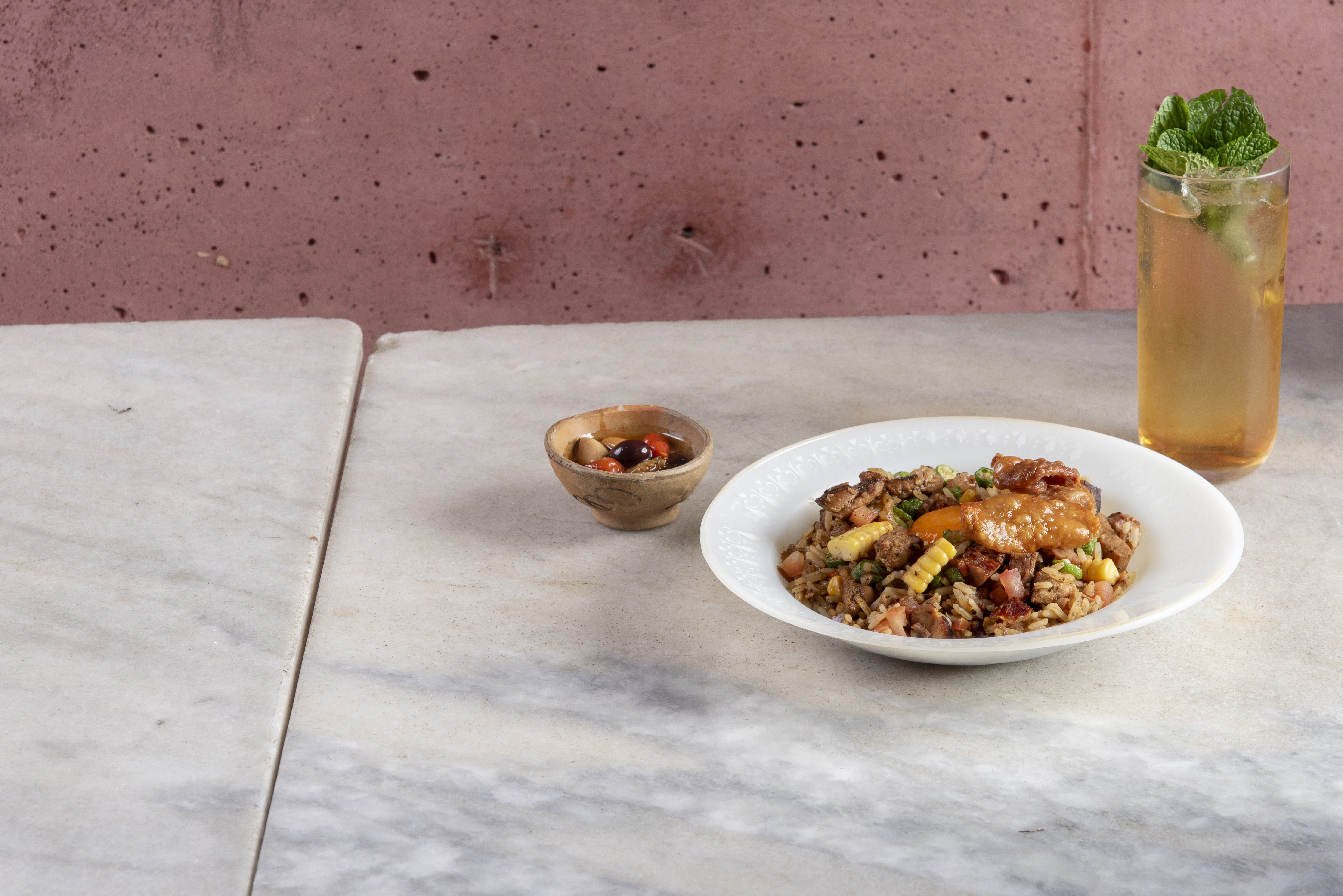 Prato de arroz frito com carne, milho e molho, acompanhado por uma tigela pequena de azeitonas e um copo de bebida com hortel&atilde;, sobre uma mesa de m&aacute;rmore branco e fundo rosa texturizado