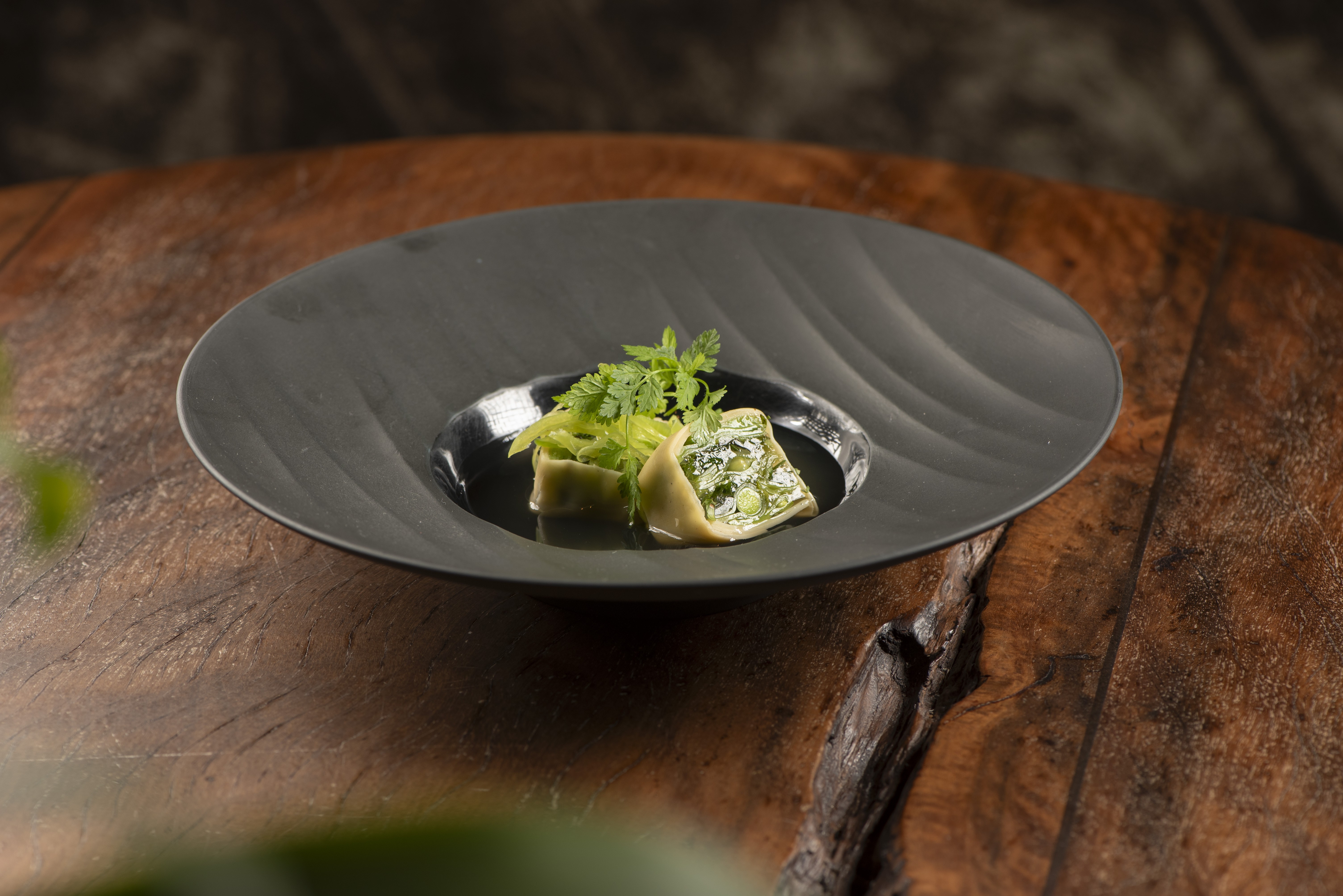 Prato gourmet com dois ravi&oacute;lis verdes, um aberto mostrando o recheio, guarnecidos com talos de aipo e folhas de salsinha, sobre um prato preto com borda ondulada, em uma mesa de madeira r&uacute;stica