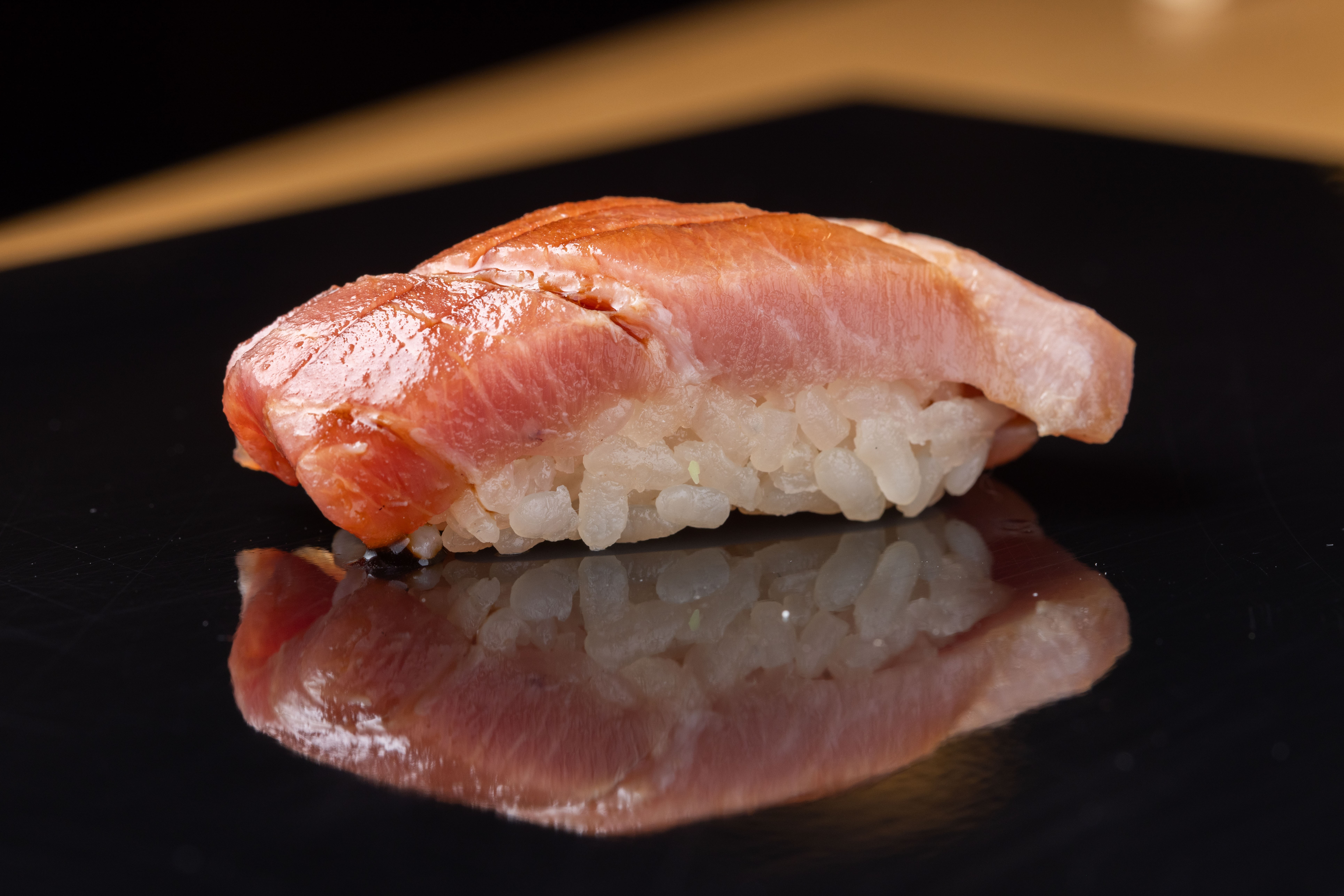 Um nigiri de atum com arroz branco, refletido em uma superf&iacute;cie preta brilhante, com um fundo escuro e uma faixa marrom desfocada