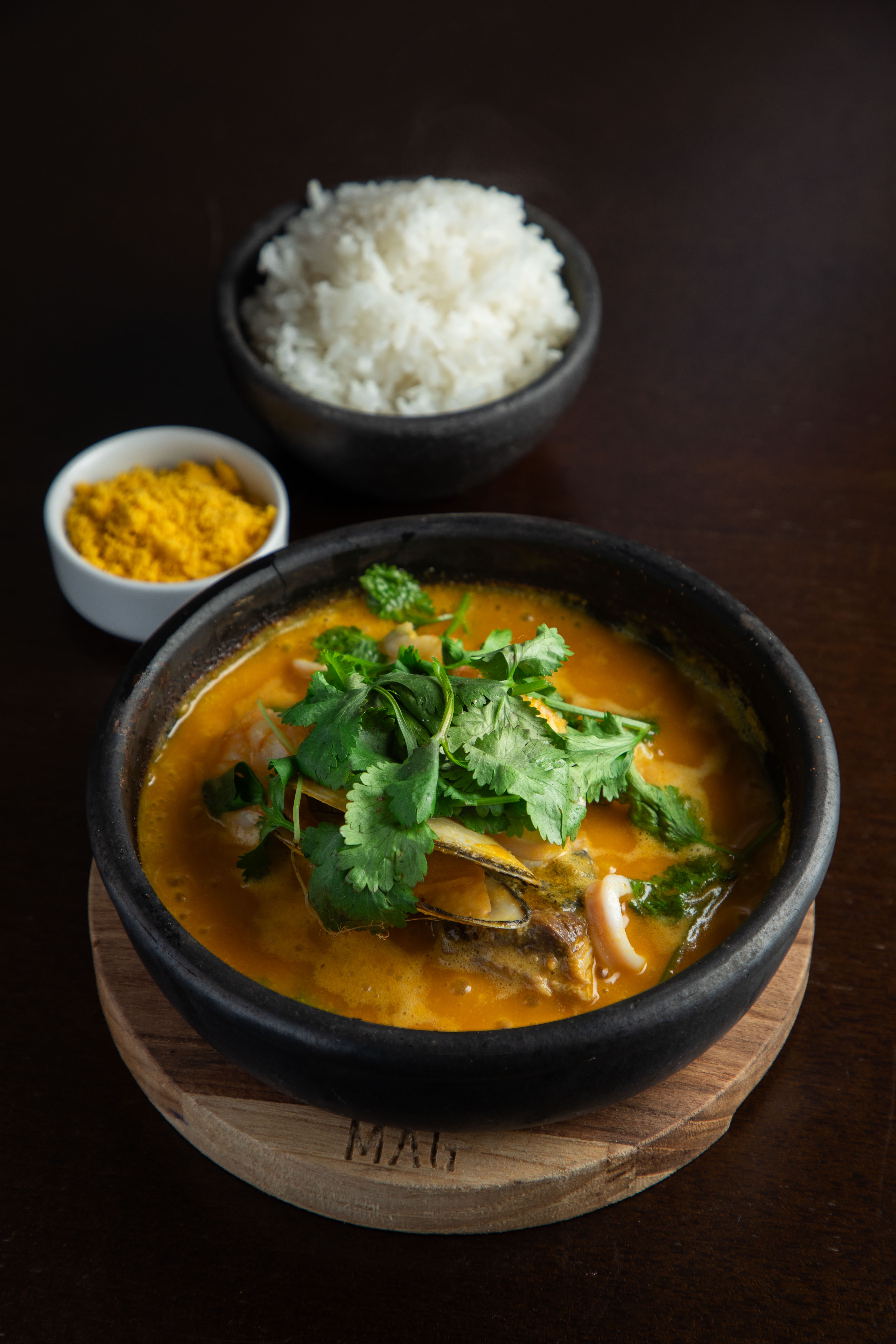 Moqueca de frutos do mar em panela de barro preta, com camar&atilde;o, lula e mexilh&otilde;es, guarnecida com coentro fresco. Ao lado, uma tigela de arroz branco e outra de farofa amarela, sobre uma mesa de madeira escura