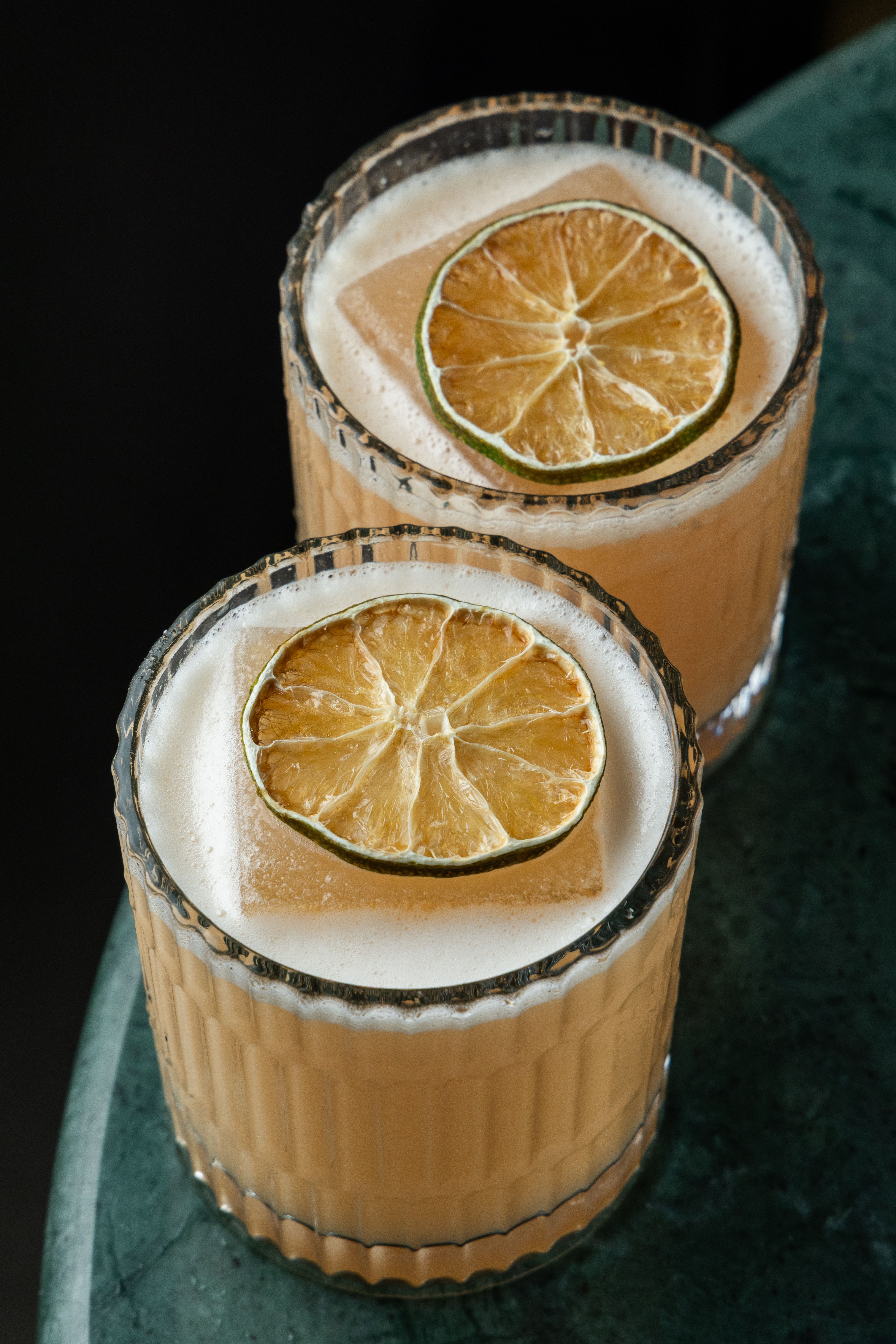 Dois coquet&eacute;is em copos texturizados, com l&iacute;quido alaranjado, espuma branca e uma rodela de lim&atilde;o desidratado sobre um cubo de gelo, em uma superf&iacute;cie de m&aacute;rmore verde escuro