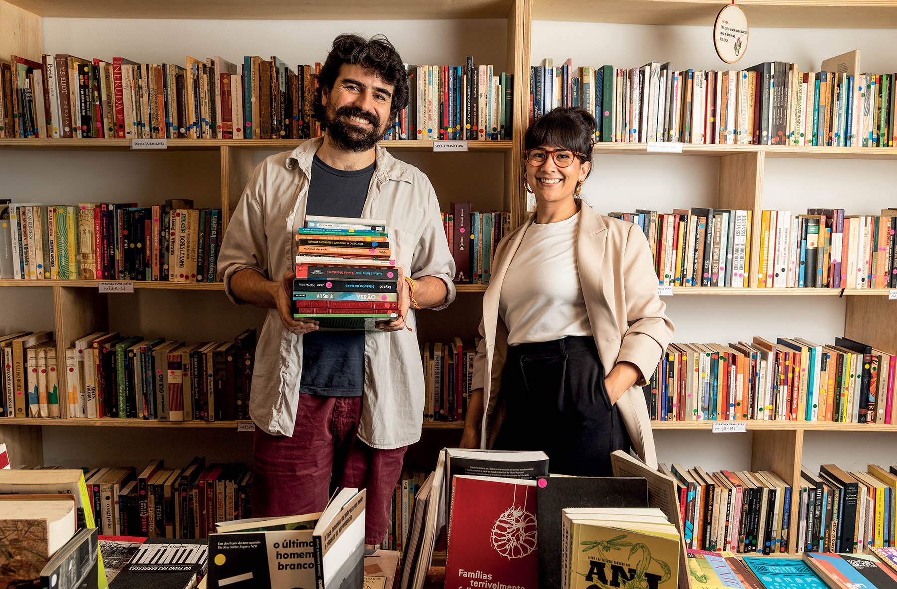 Um homem barbudo sorrindo, segurando uma pilha de livros, e uma mulher sorridente de óculos, com as mãos nos bolsos, em frente a estantes cheias de livros