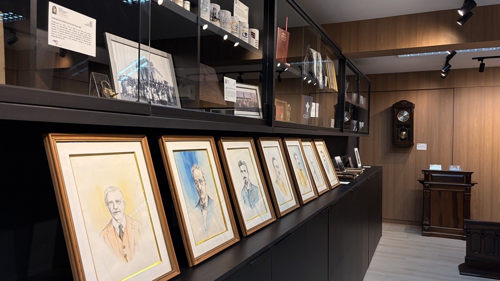 Exposição com uma fileira de sete retratos masculinos em aquarela, emoldurados, inclinados sobre um balcão escuro. Acima, prateleiras de vidro exibem documentos e objetos históricos. Ao fundo, parede de madeira com relógio antigo e móvel de madeira escura.