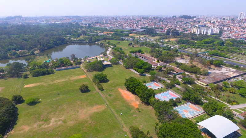Vista aérea de um parque com lago, áreas verdes e quadras esportivas coloridas, com a cidade ao fundo sob céu claro